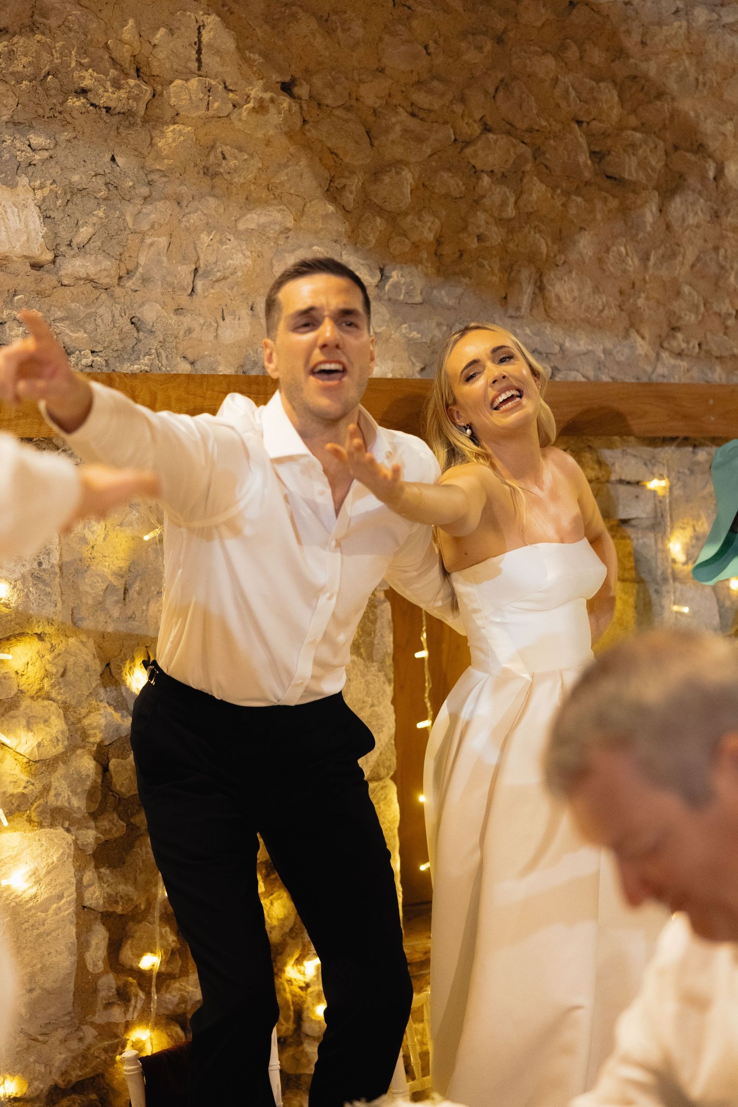 Wedding guests dance and celebrate in front of rustic stone wall with twinkle lights.