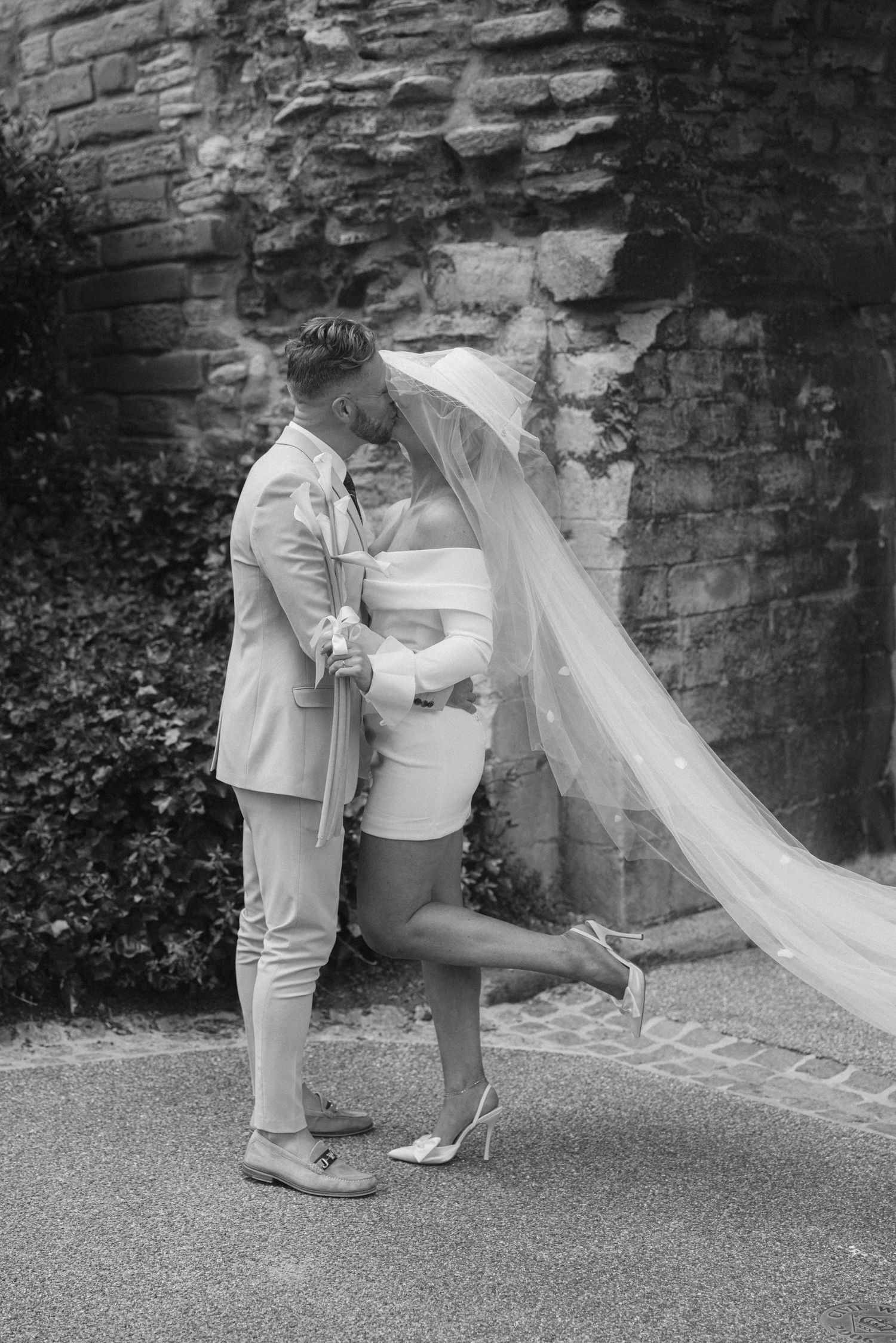 A romantic black and white portrait of newlyweds embracing against a stone wall.