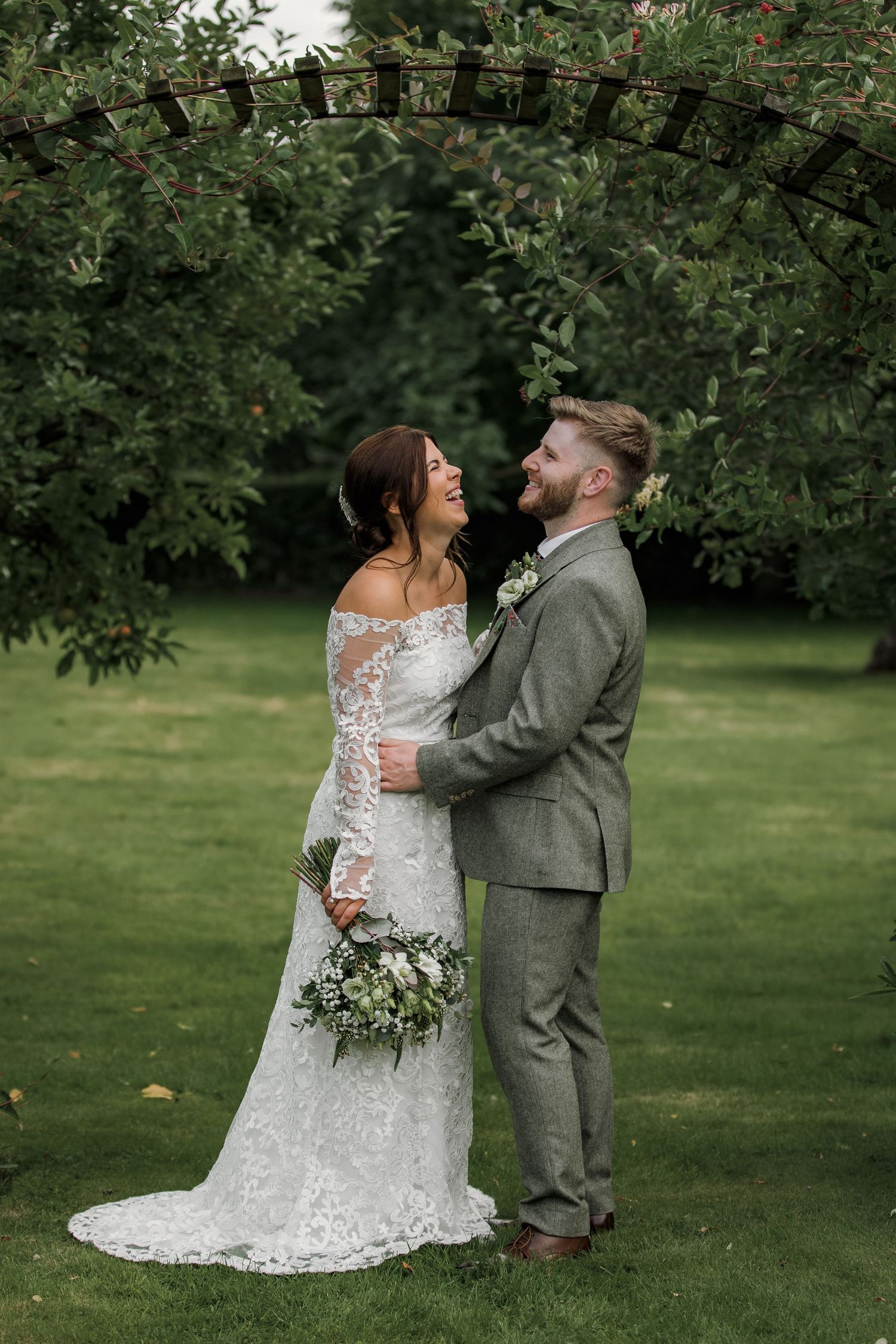A romantic wedding photo of a couple laughing together under a garden arch with lush greenery.
