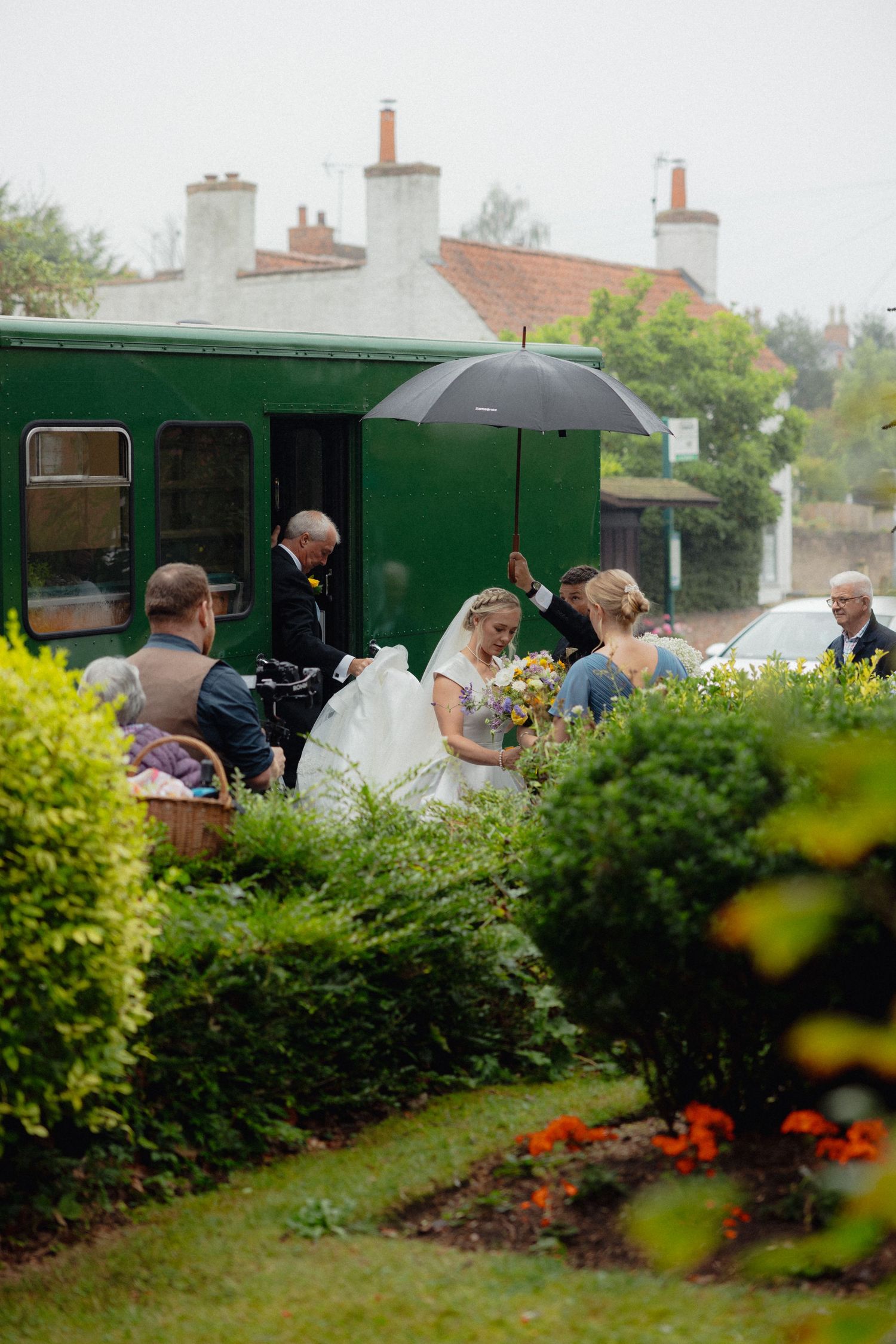 Wedding party gathered beside a vintage green train car surrounded by lush garden landscaping.