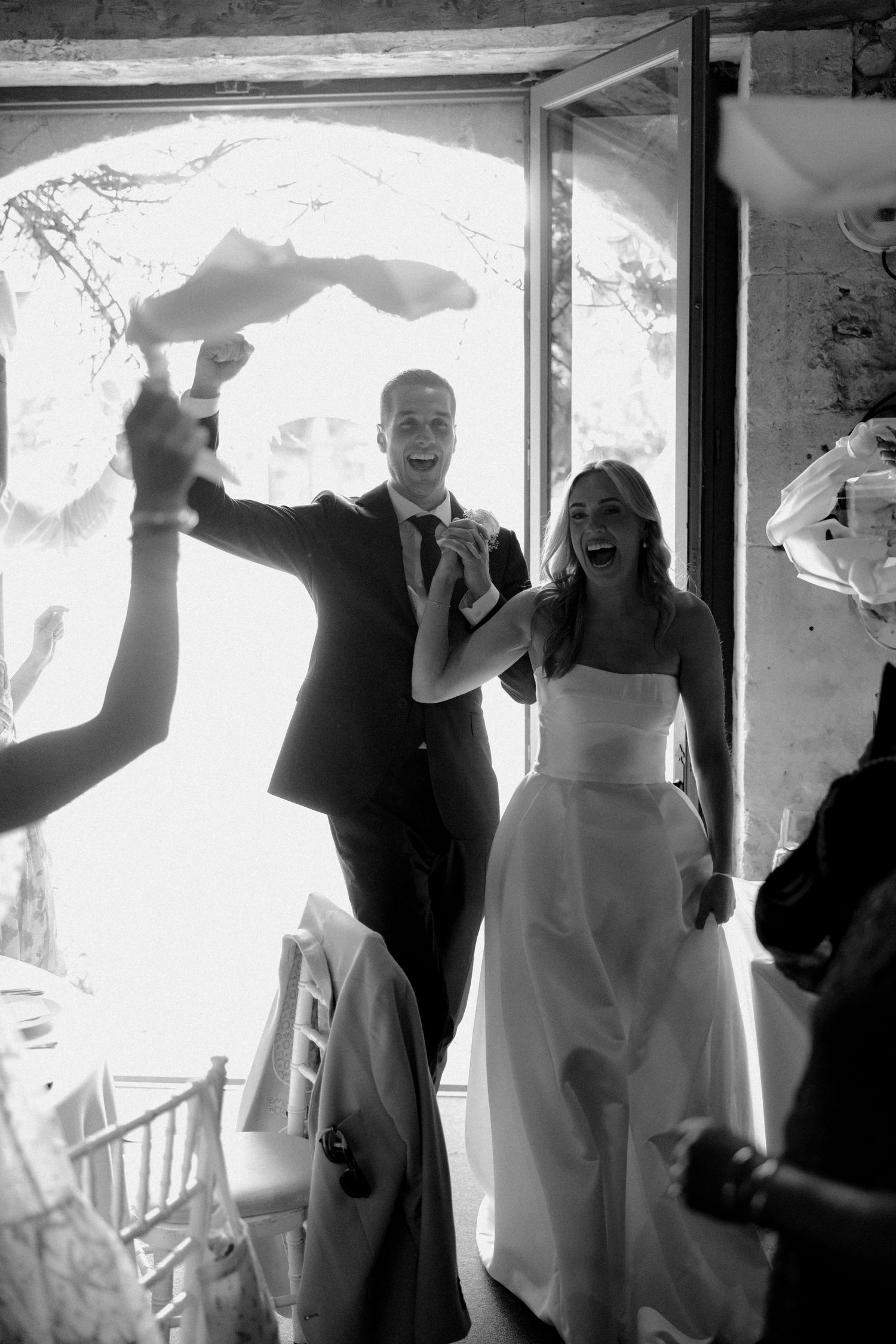 Wedding party celebrates with joyful dancing in front of bright windows.