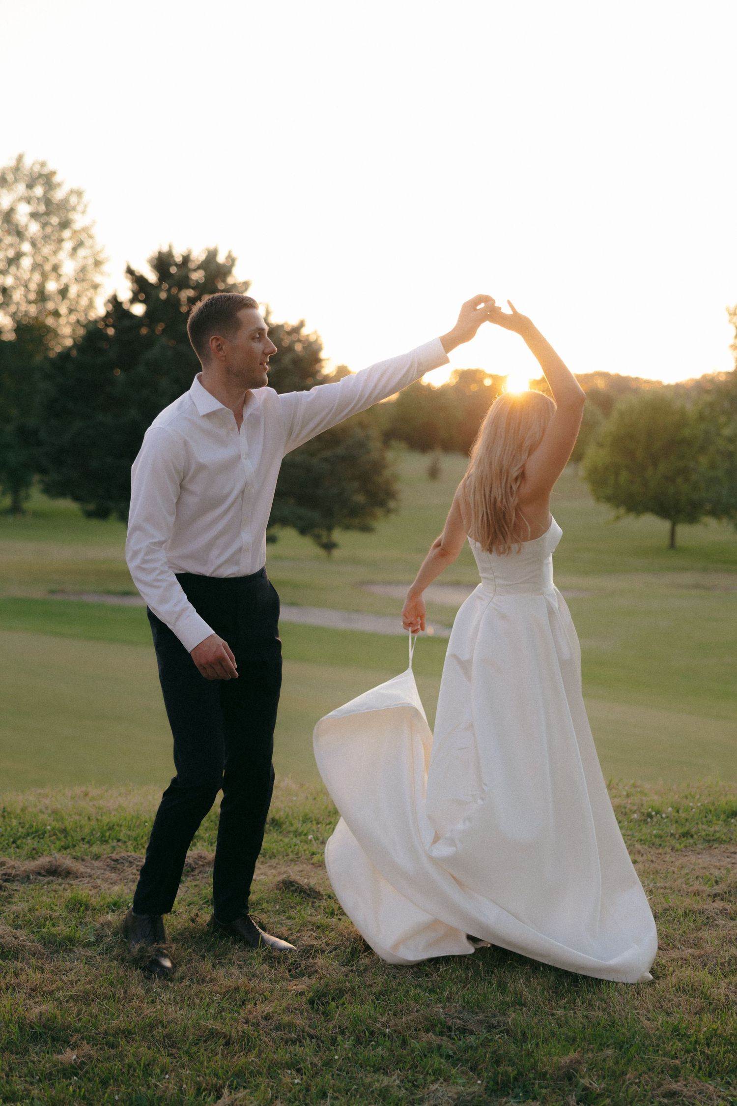 Newlyweds dance playfully in a grassy field during golden hour.