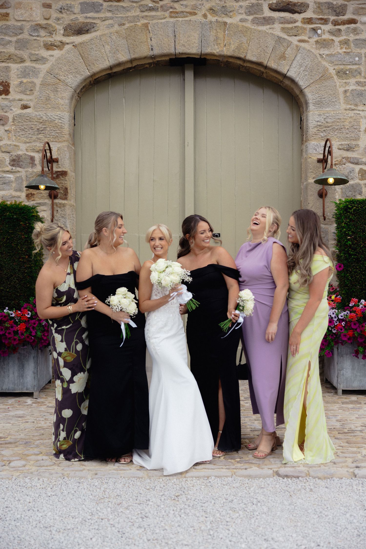 Wedding party laughing together under stone archway with colorful flowers.