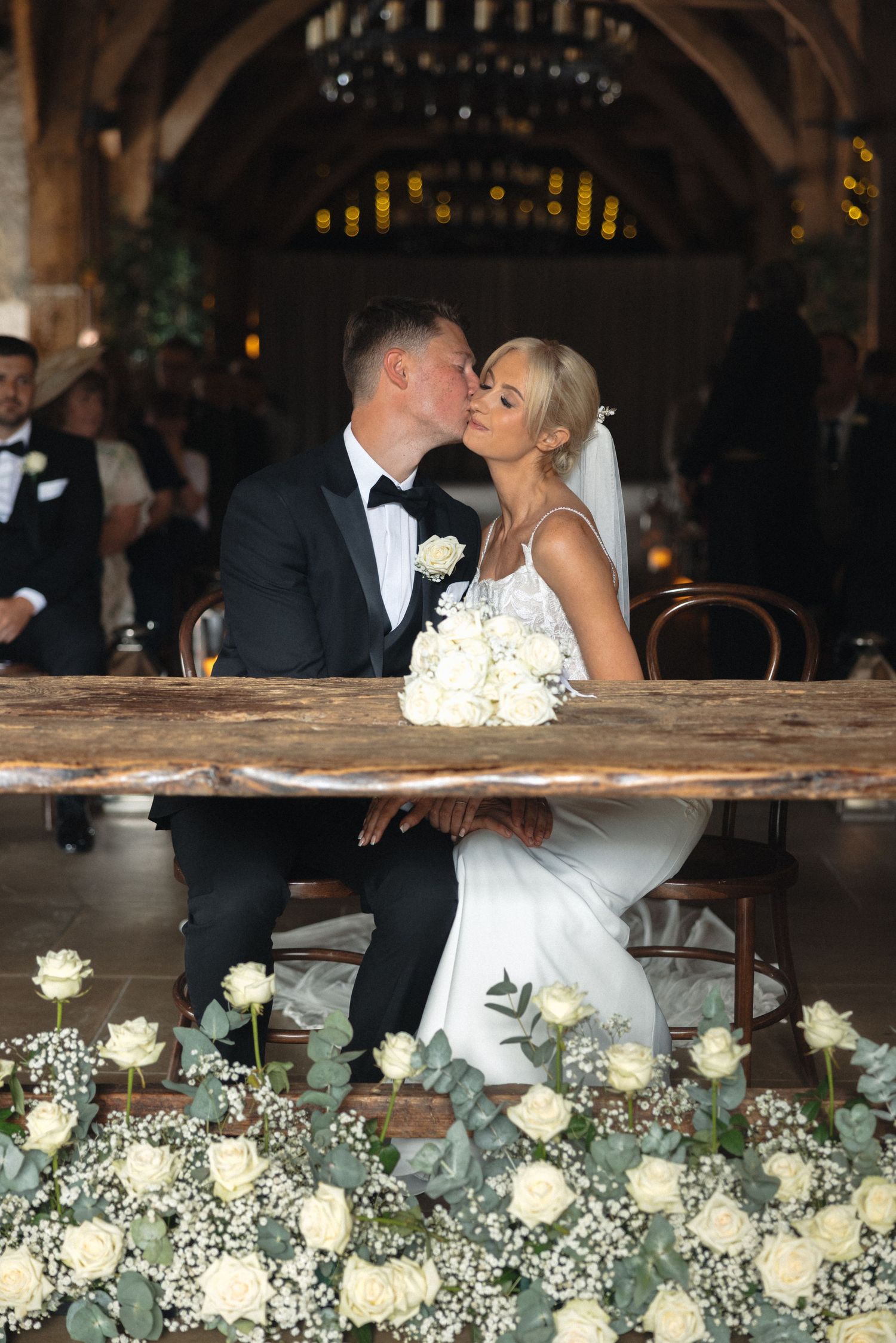 Wedding reception decor featuring white roses and baby's breath arrangements along rustic wooden surfaces.