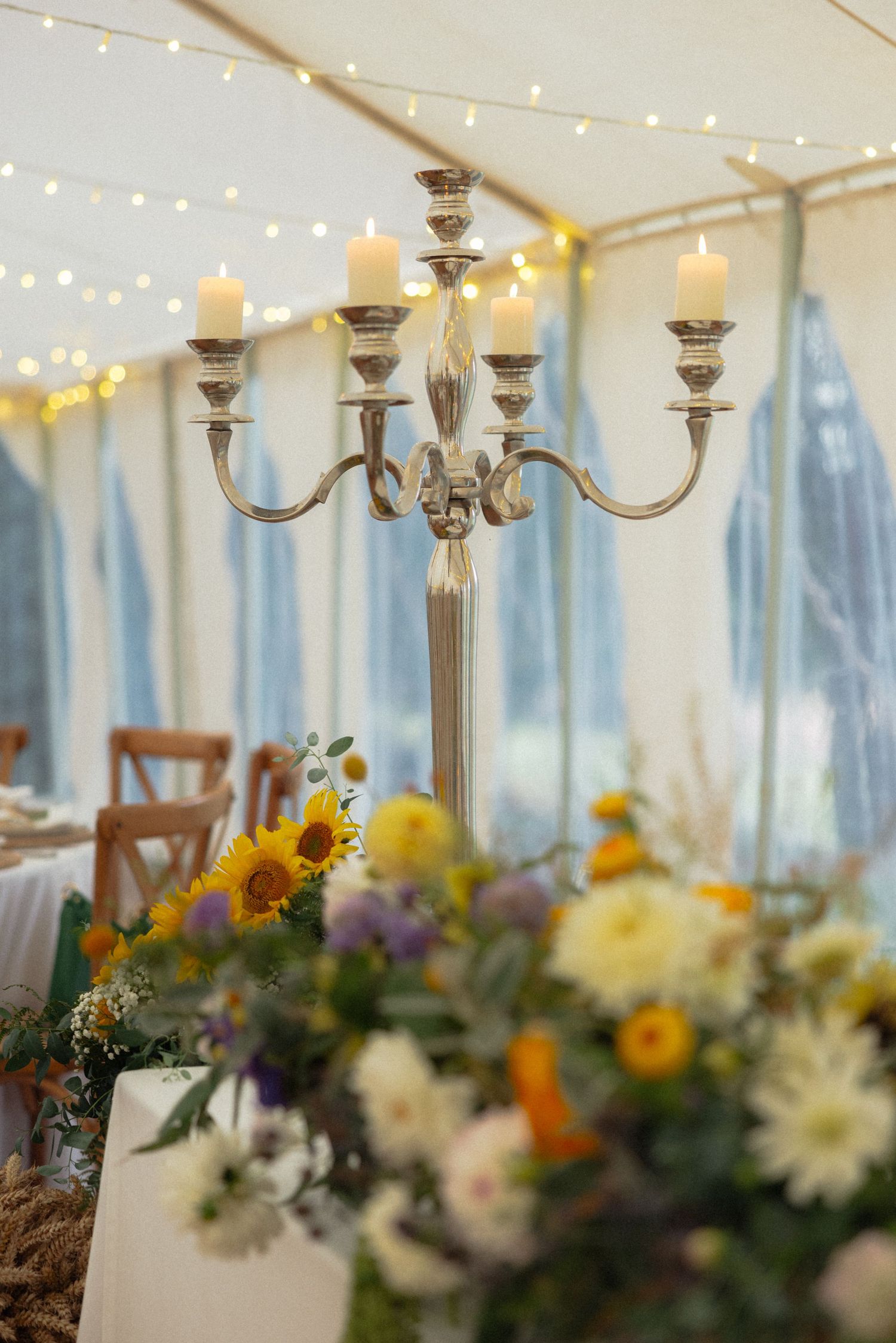 Elegant candelabras and floral arrangements line wedding reception tables under a white tent with string lights.