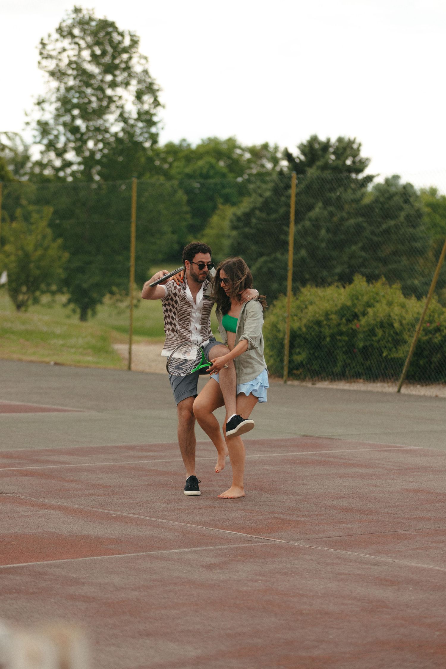 A couple dancing and playing around on an outdoor tennis court.