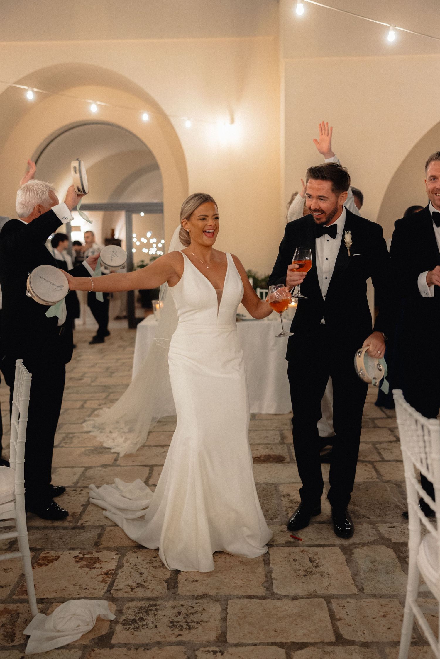 Wedding party dances enthusiastically on a stone floor under arched ceilings with string lights.