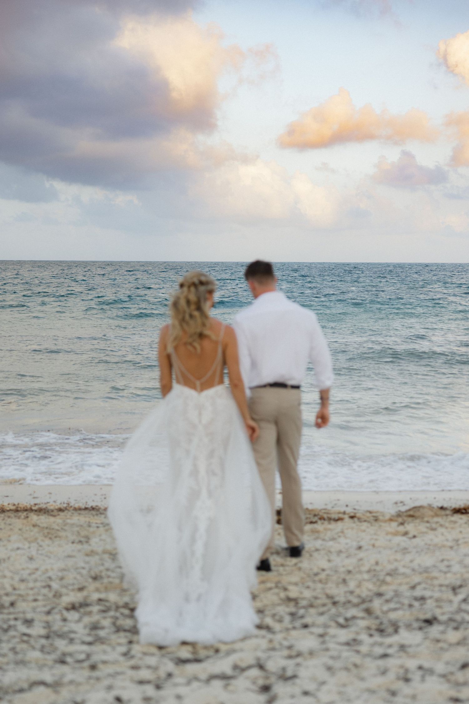 Couple walks along a tropical beach at sunset in wedding attire with waves rolling in.