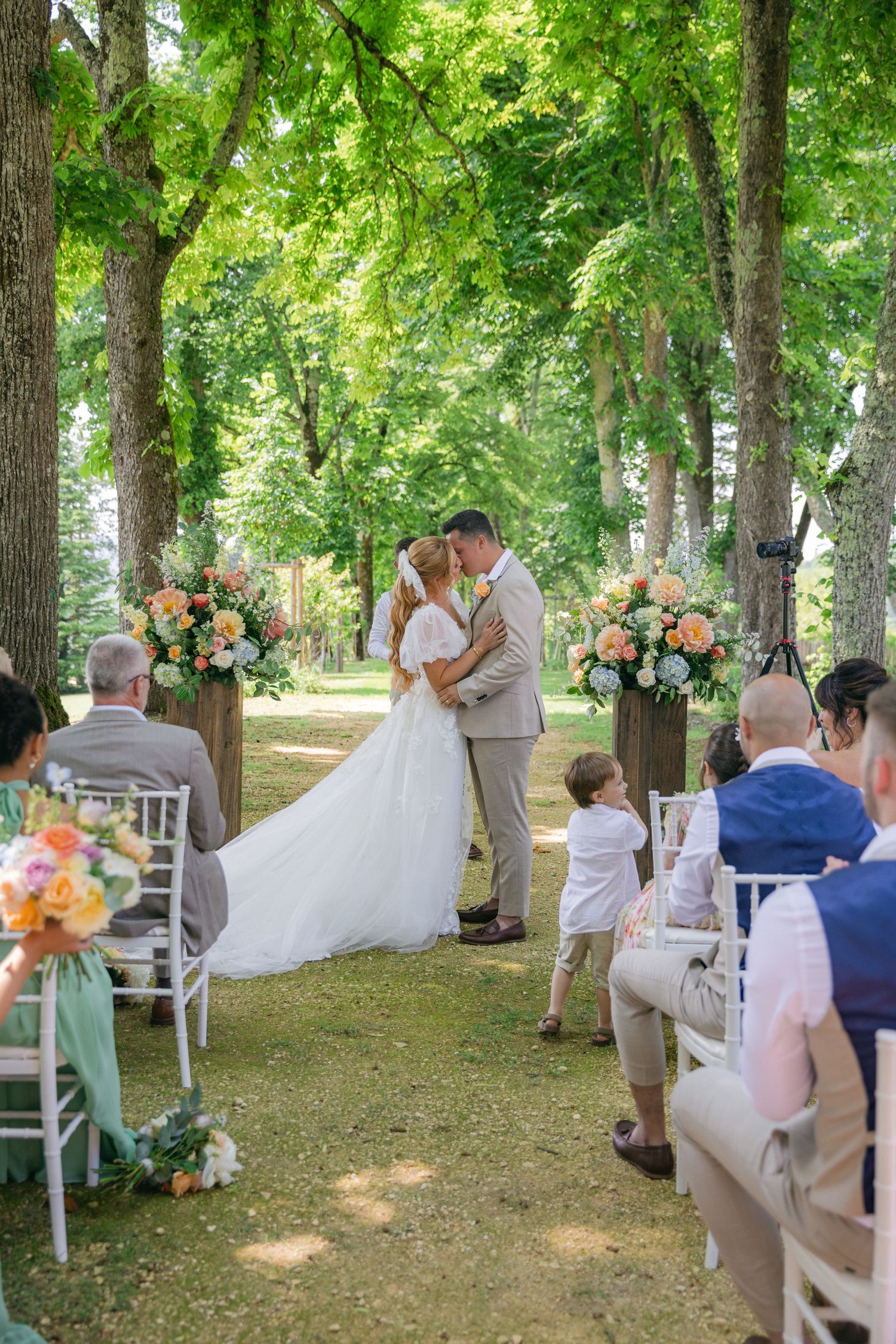 An outdoor wedding ceremony in a wooded area with white chairs and floral decorations.