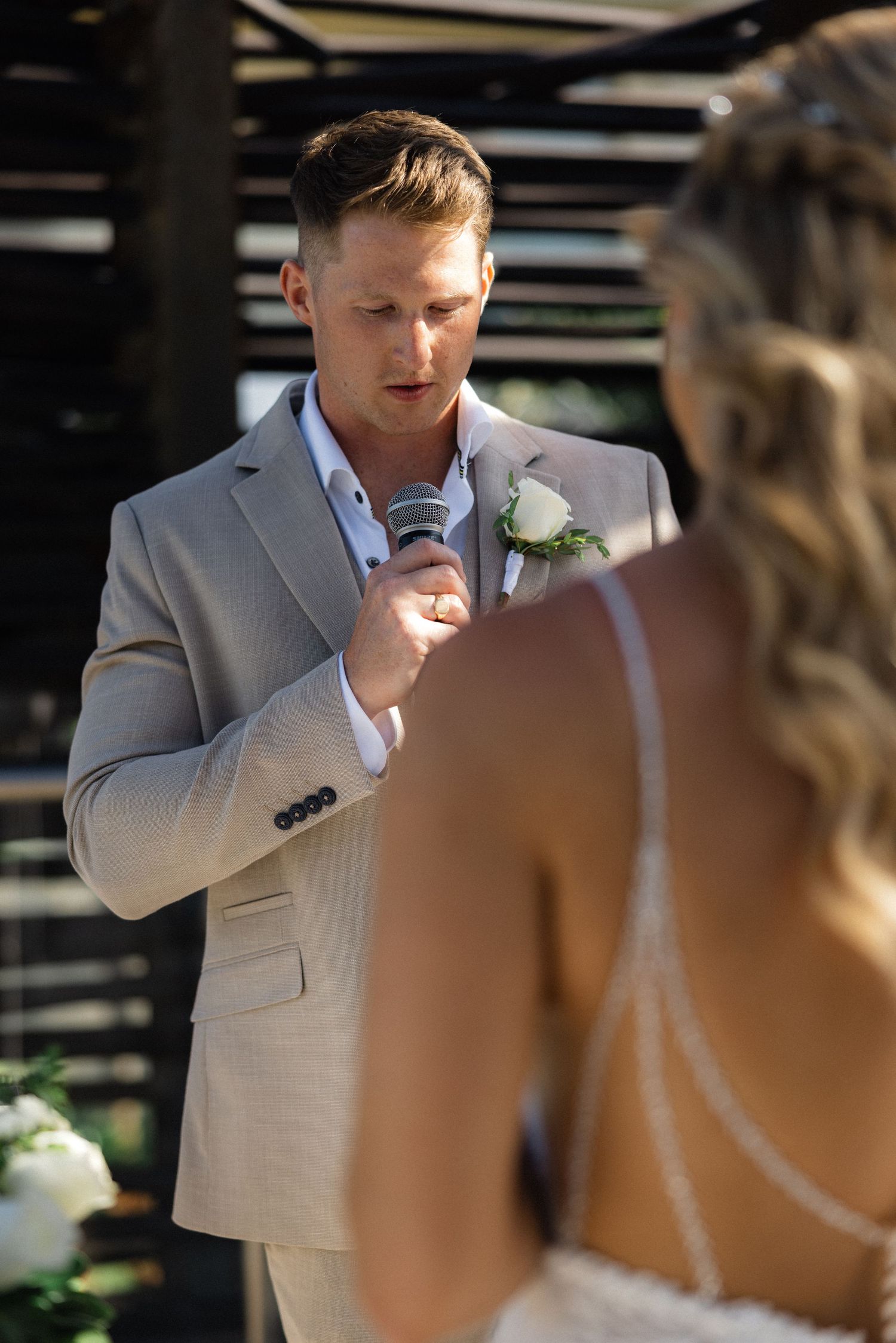 Groom in light gray suit speaks emotionally into microphone during outdoor wedding ceremony.