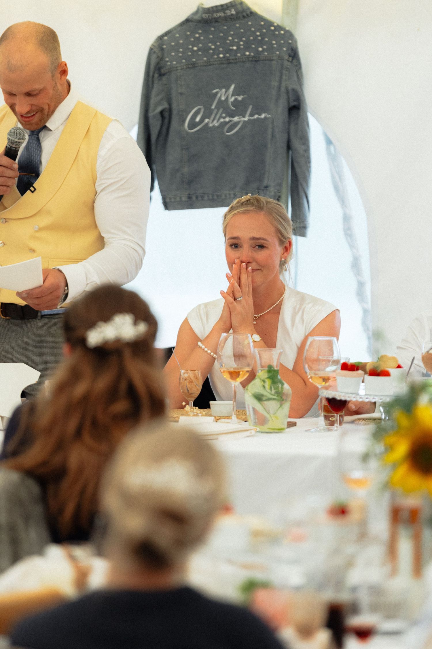 Wedding guests laugh and enjoy a meal at elegantly decorated reception tables with yellow accents and string lights.