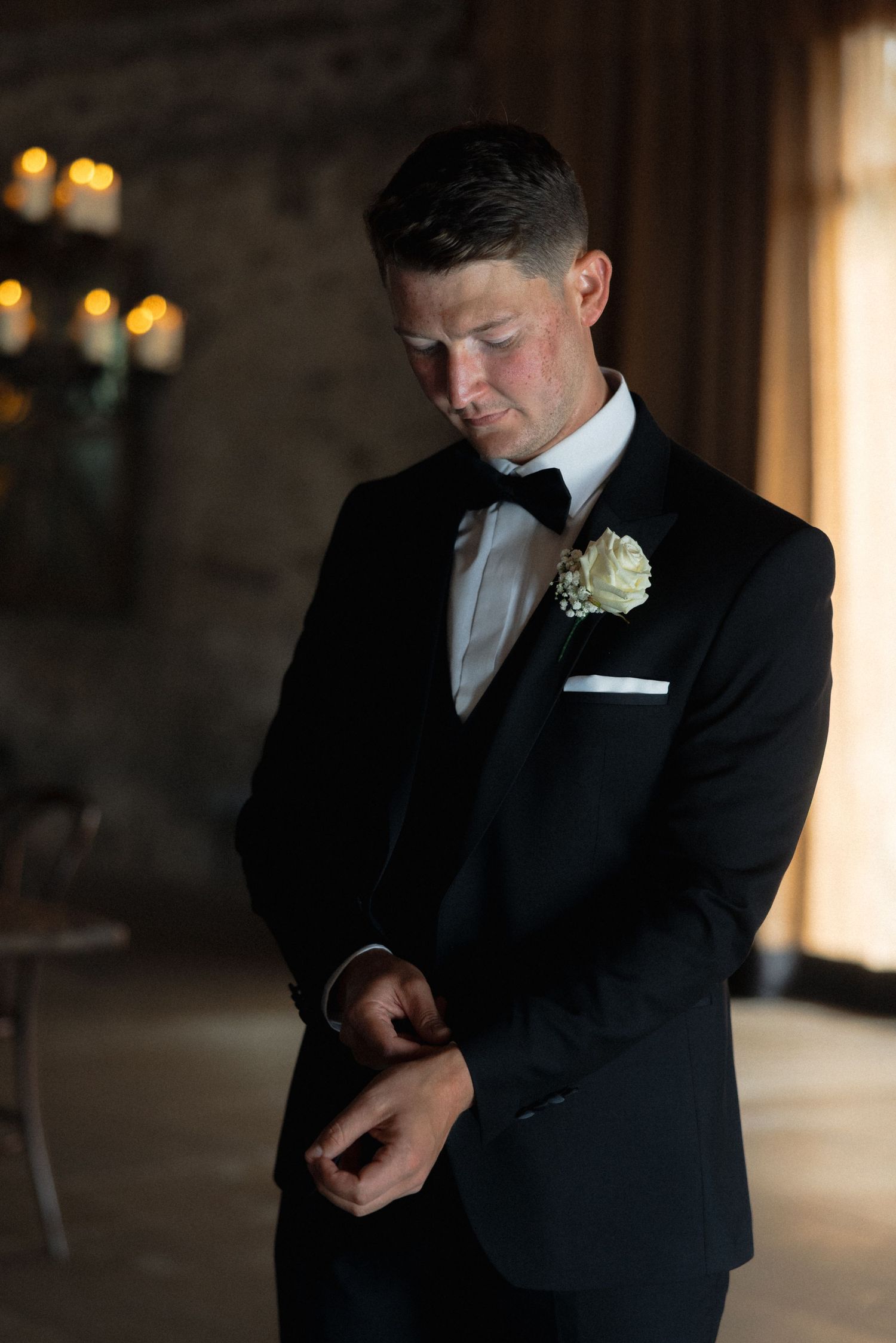 Groom in classic black tuxedo adjusting cufflinks in dimly lit venue.