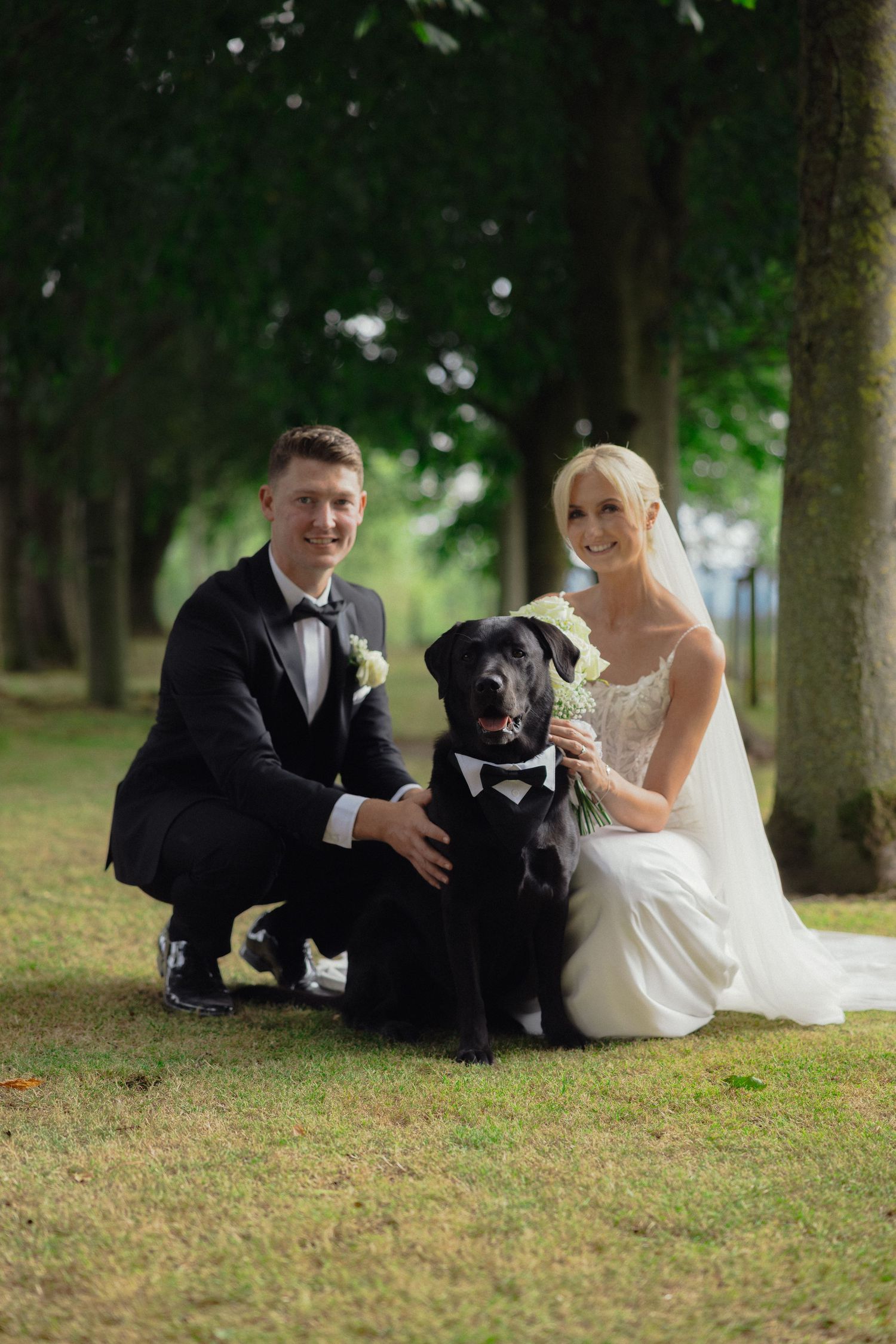 A wedding couple poses with their black dog on a grassy lawn surrounded by trees.