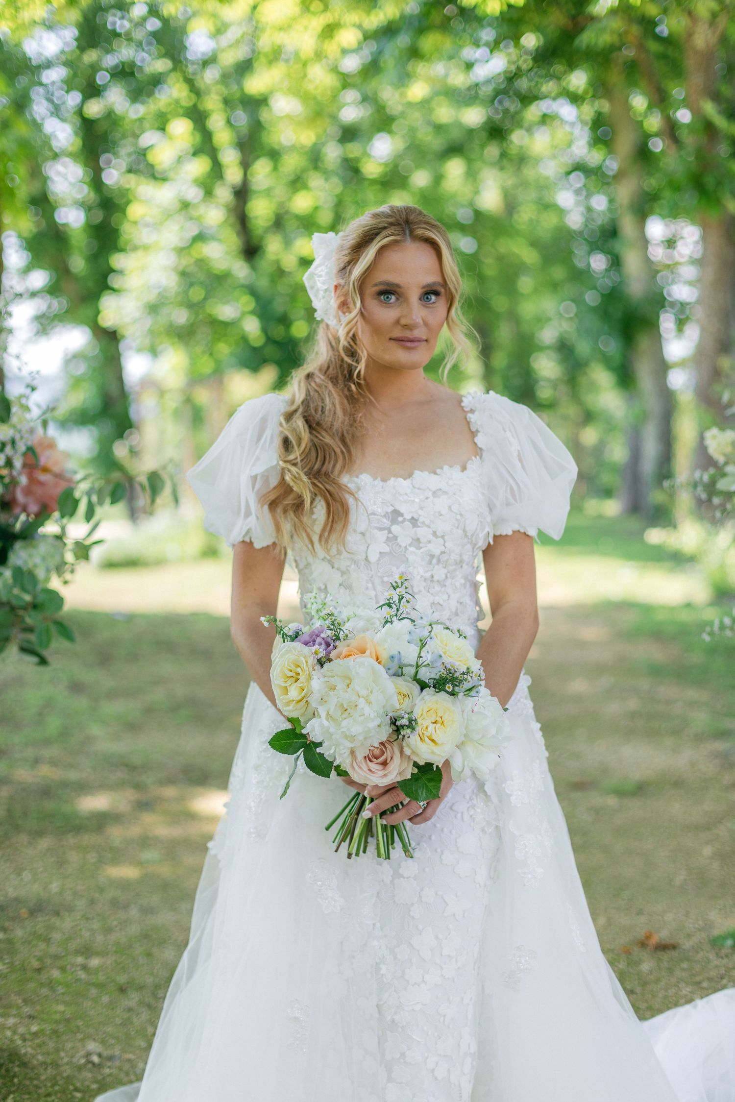 A bride in an elegant white gown poses with her bouquet in a sunlit forest setting.