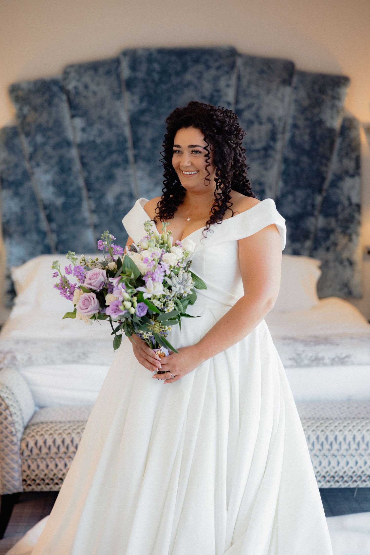 A bride in a flowing white dress poses with purple flowers against an elegant backdrop.