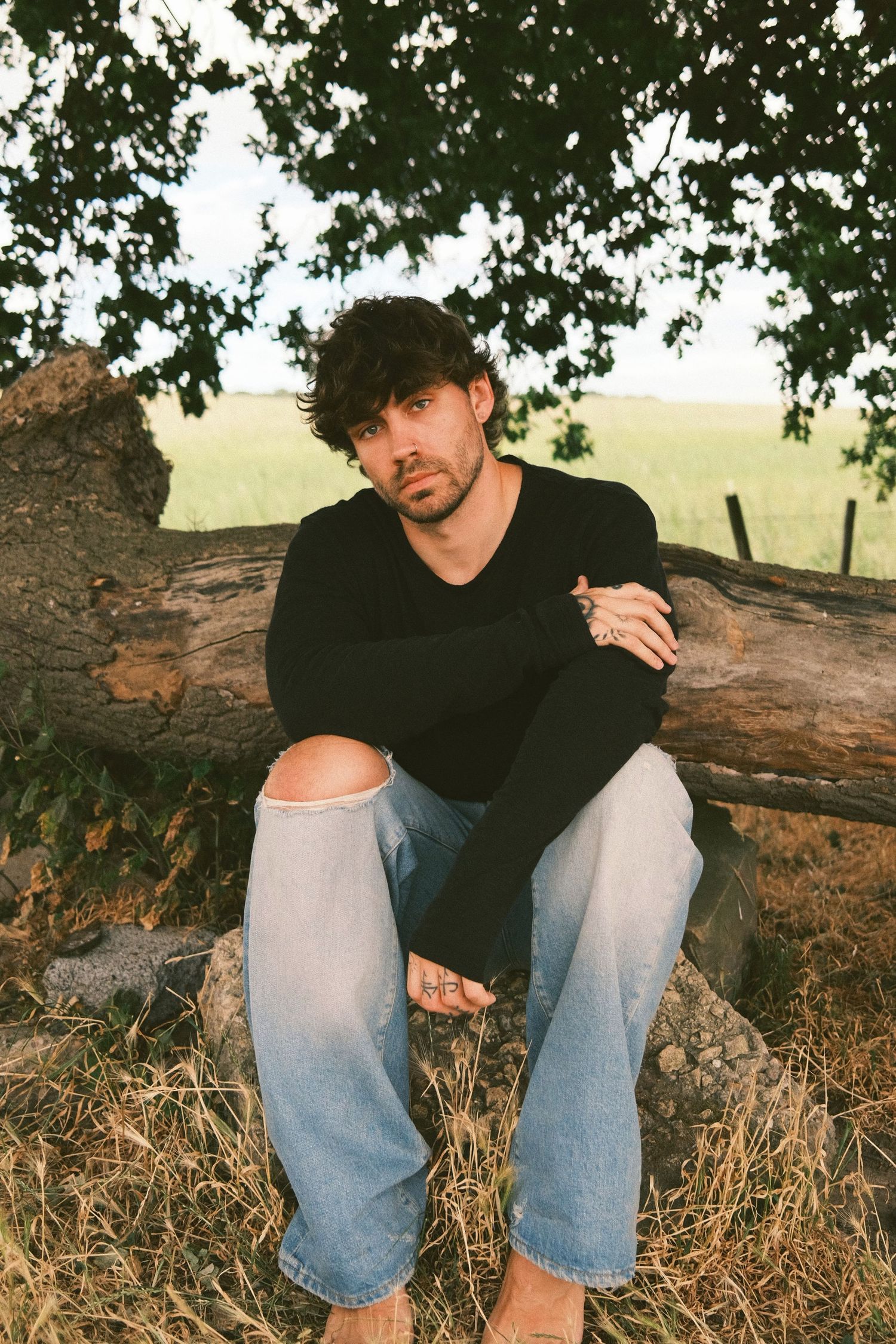 Casual portrait of man in black sweater and ripped jeans sitting on fallen tree trunk outdoors.