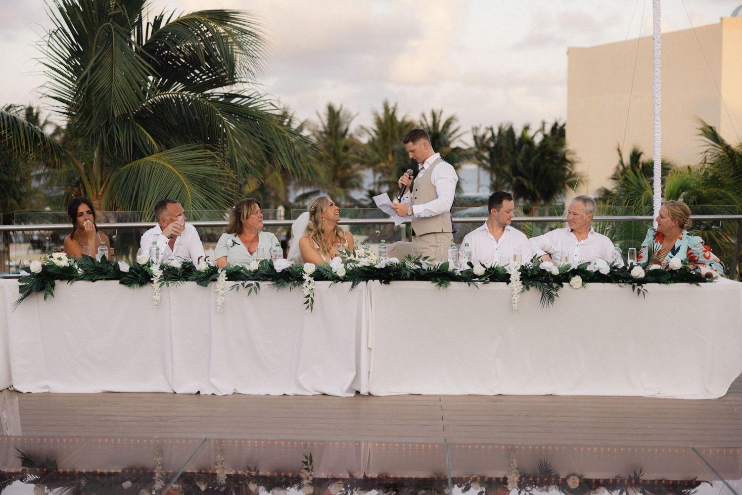Long wedding reception table setup against a tropical sunset backdrop with palm trees.