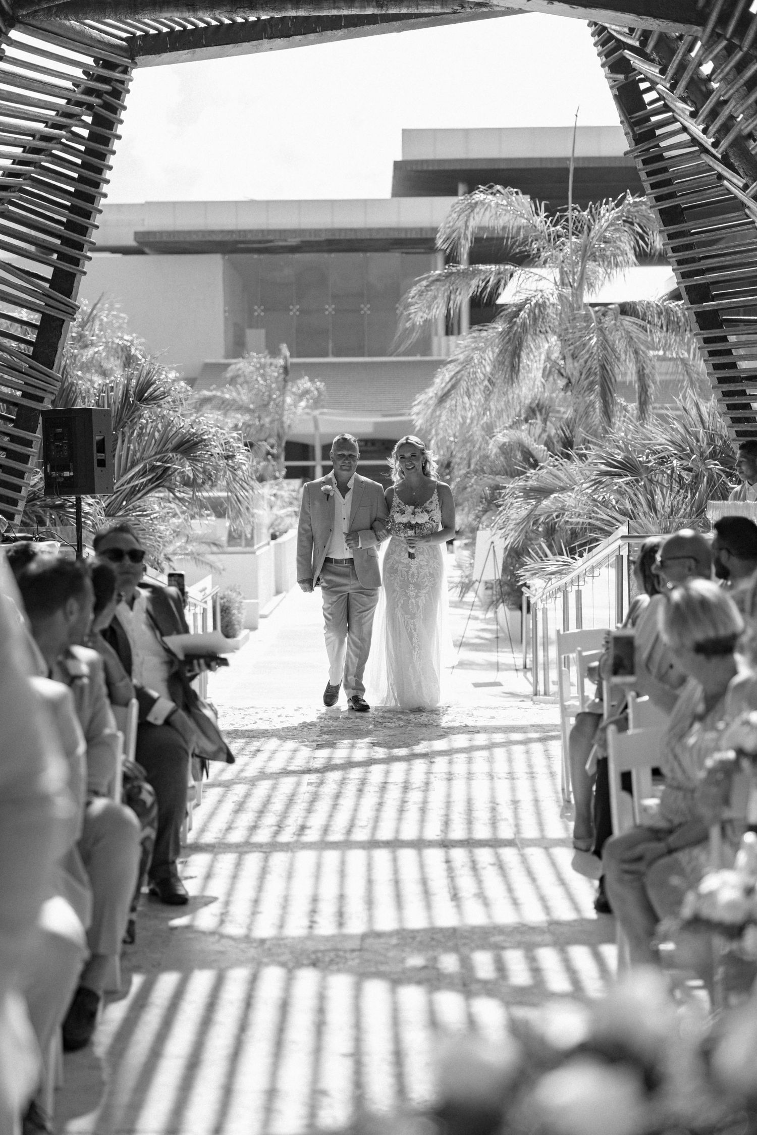 Wedding guests seated under a slatted pergola creating dramatic shadow patterns.