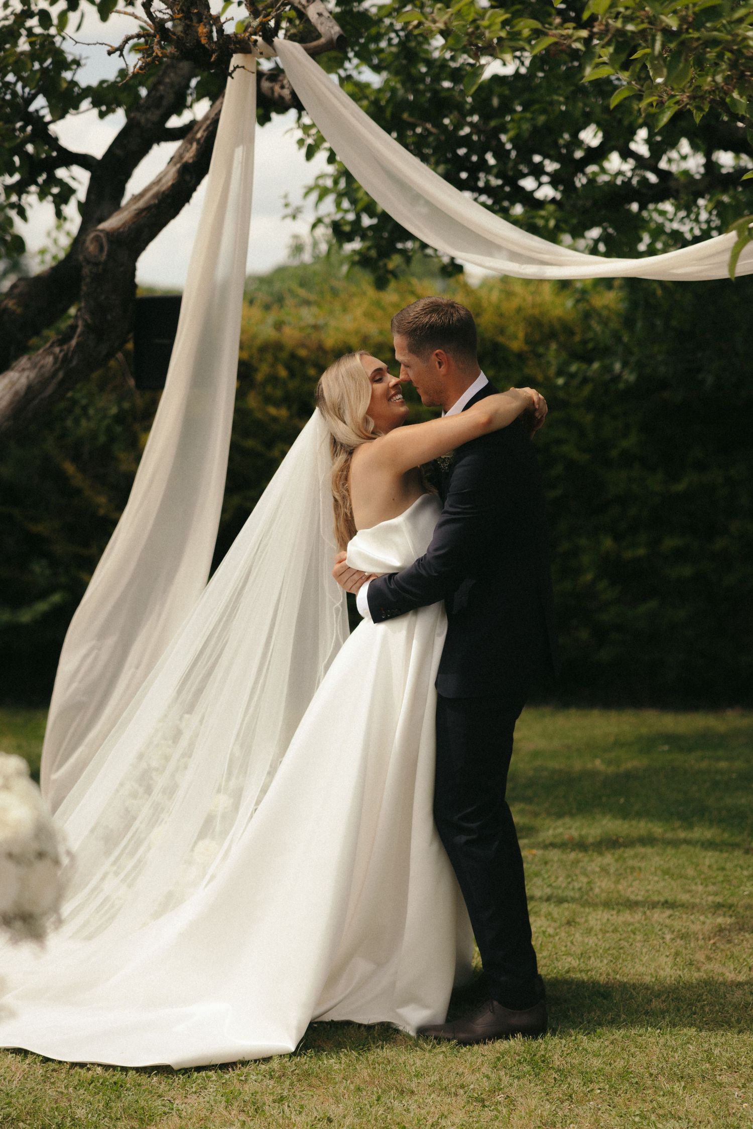 A romantic embrace under flowing white fabric during an outdoor wedding ceremony.