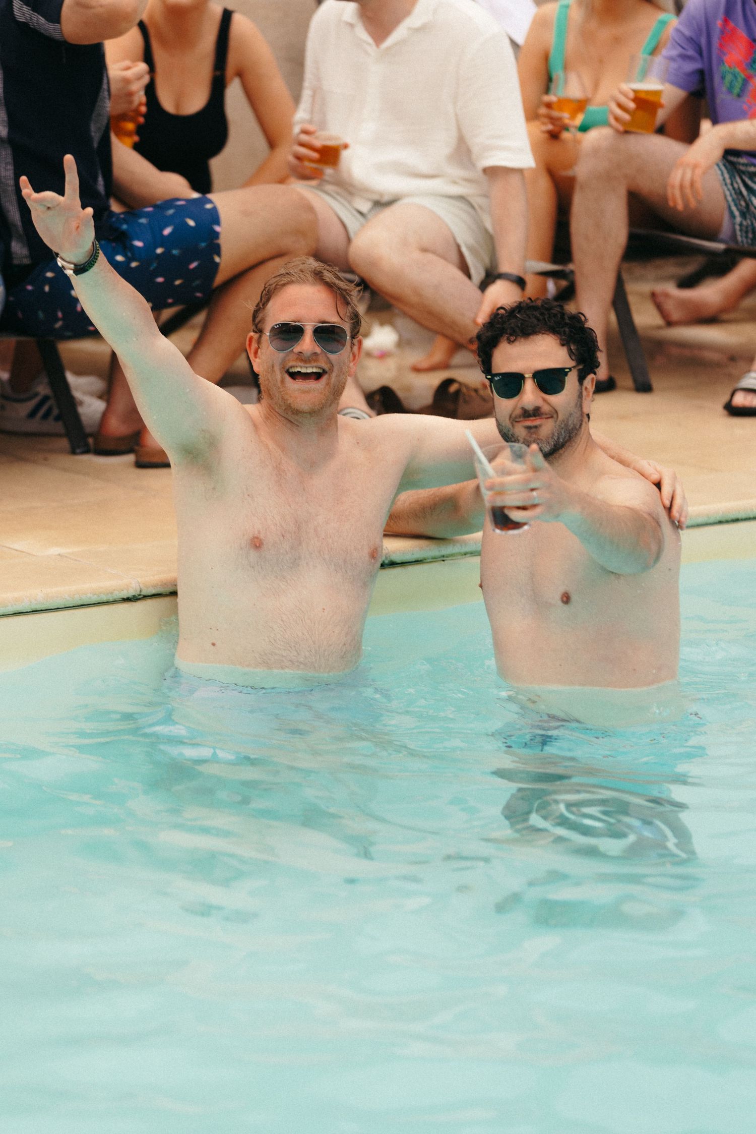 People celebrating in a swimming pool with raised arms and drinks.