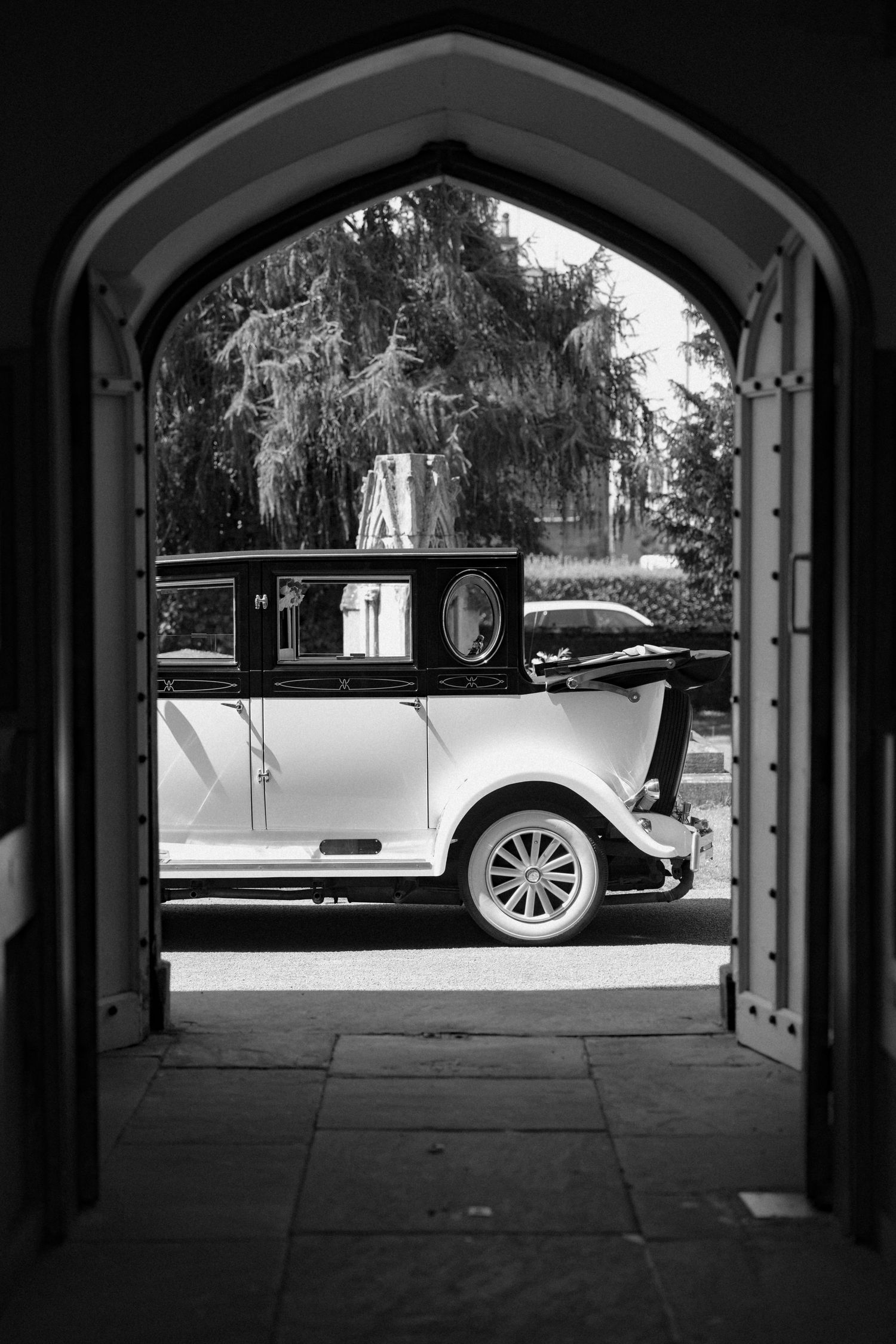 Classic black and white photo of vintage wedding cars through an arched passageway.