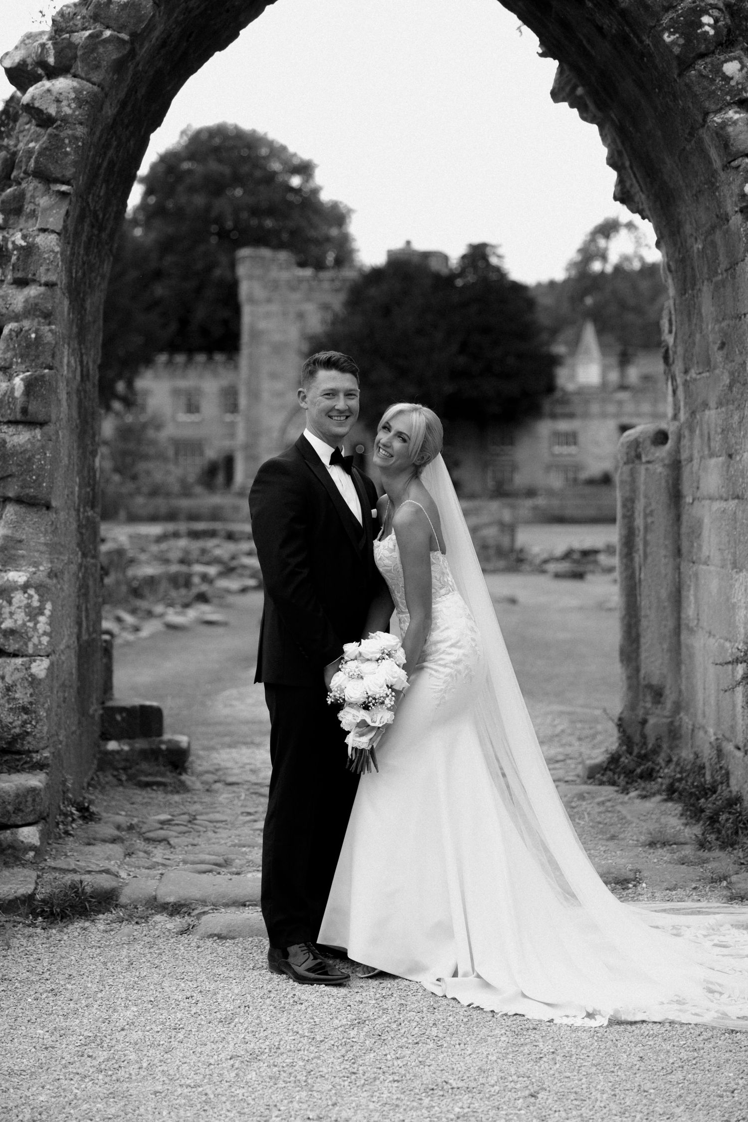 A black and white wedding portrait under an ancient stone arch with castle ruins in background.