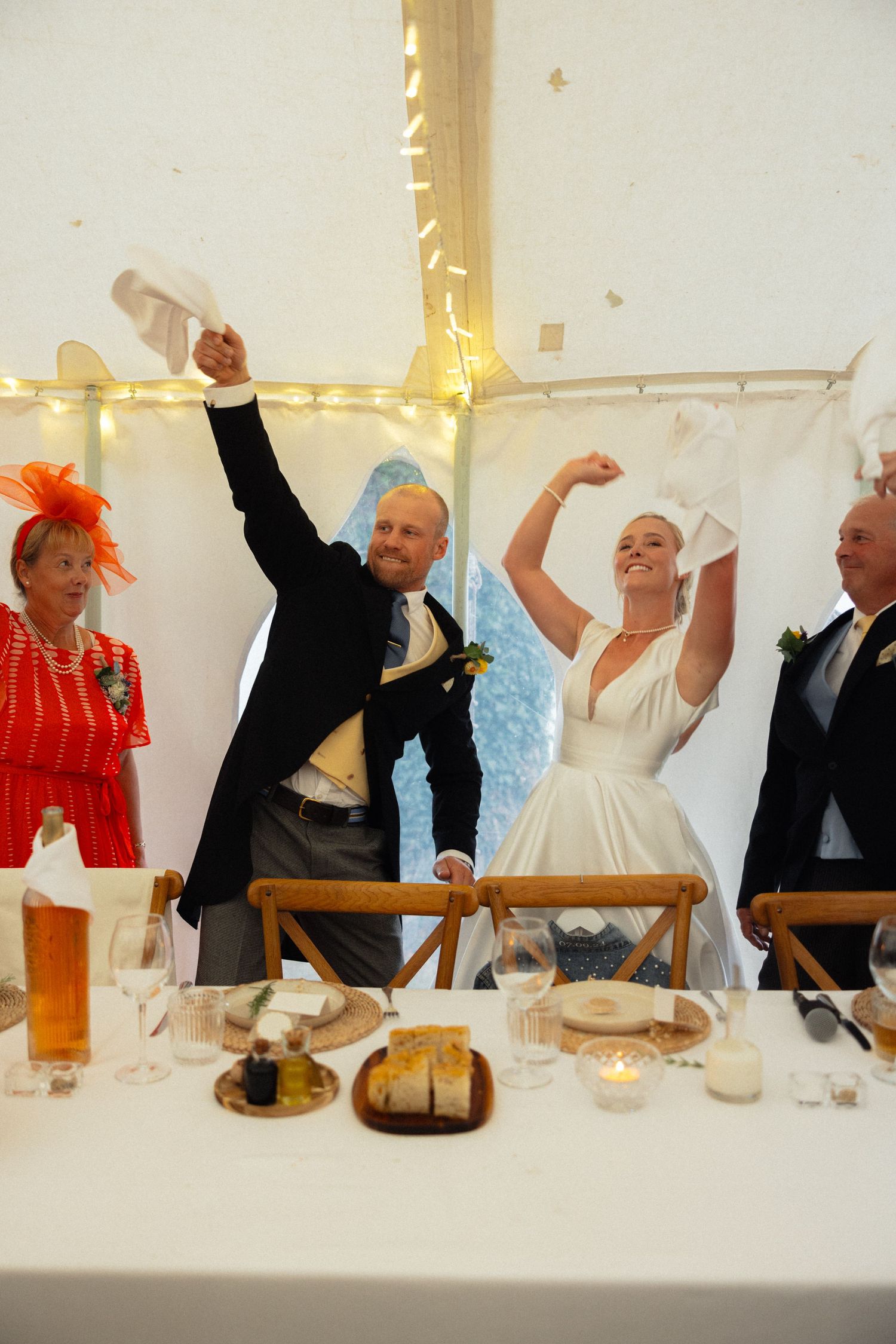 Wedding guests celebrate joyfully at a reception table decorated with string lights in a tent.