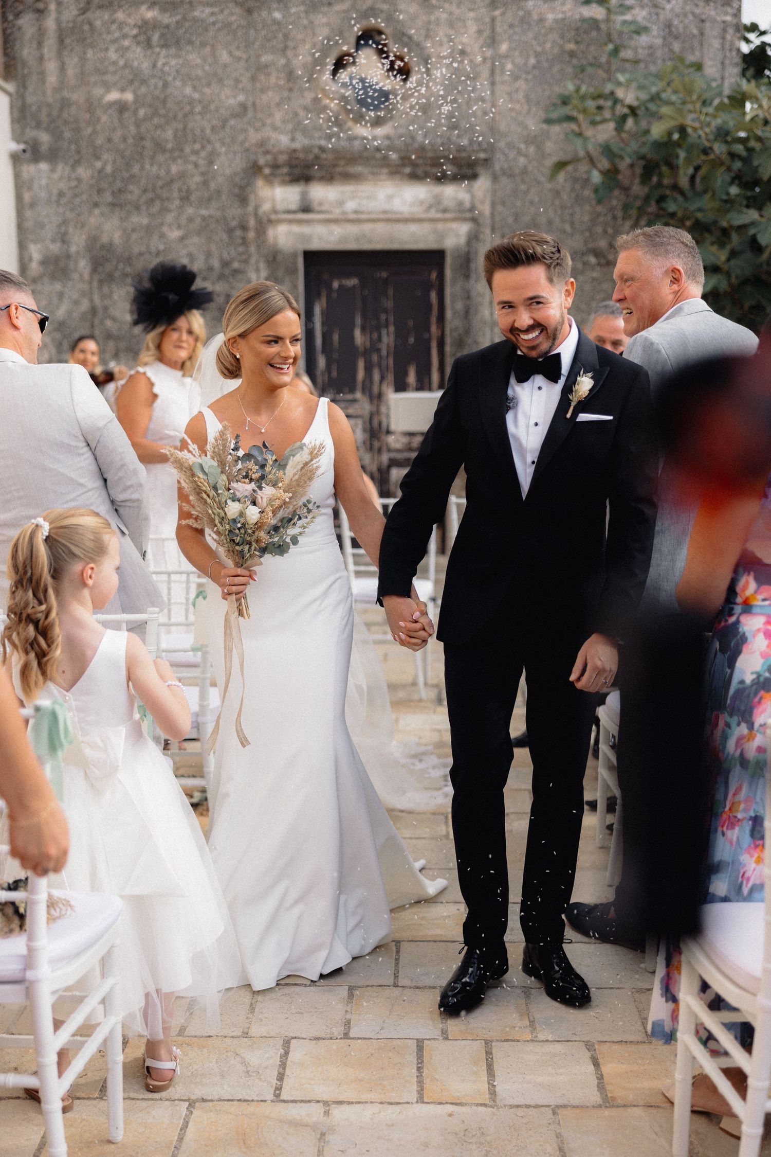 Wedding celebration with guests and couple on stone patio with white chairs.