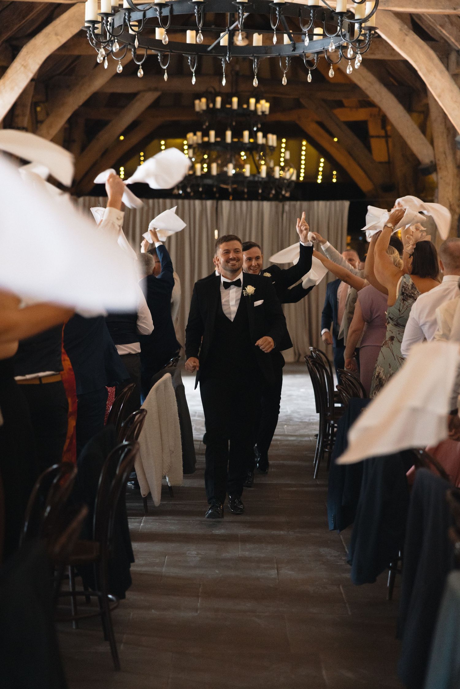 Wedding guests wave napkins during a celebratory entrance in a rustic venue.