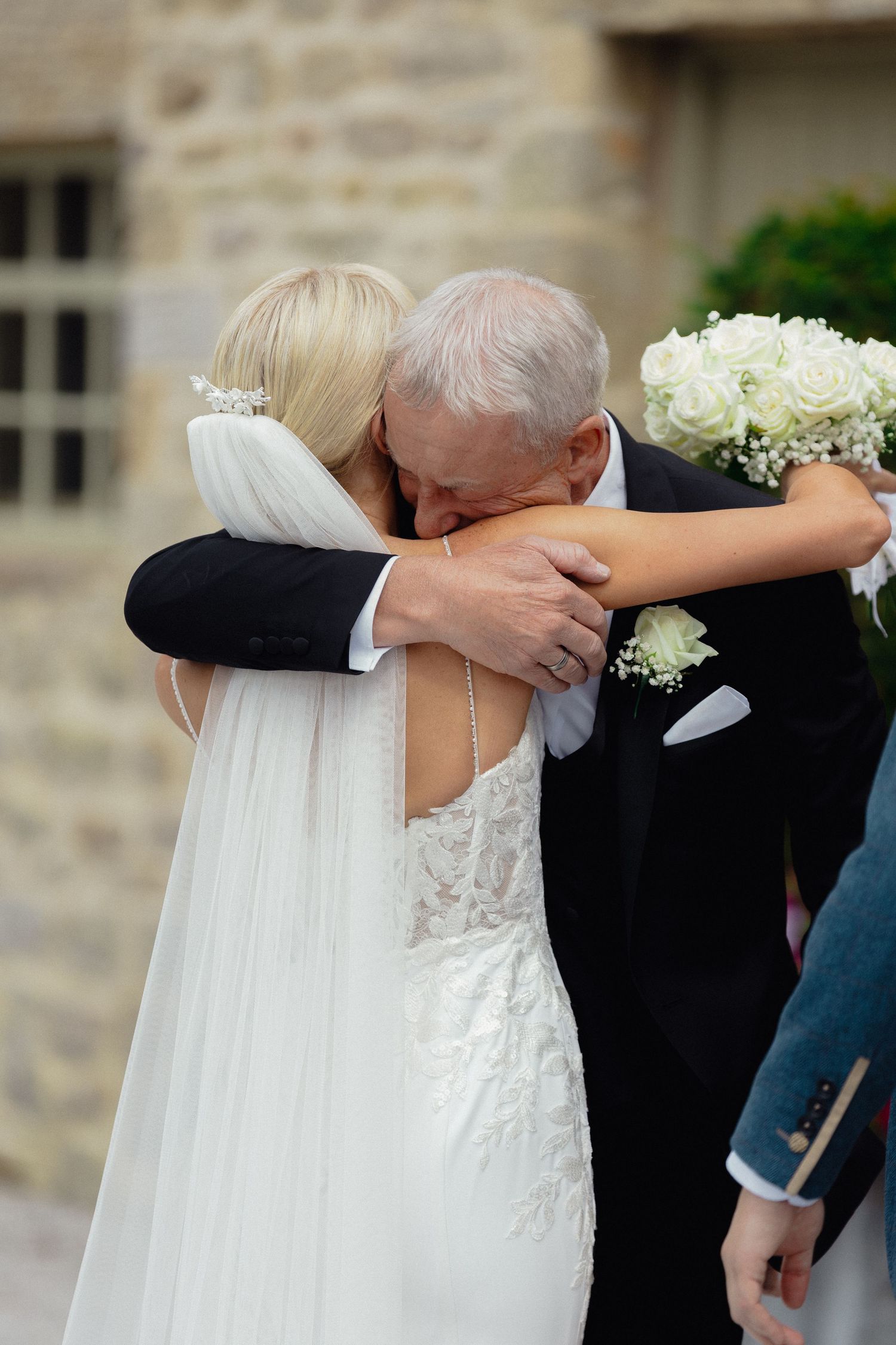 An emotional embrace between wedding participants outside a stone building.