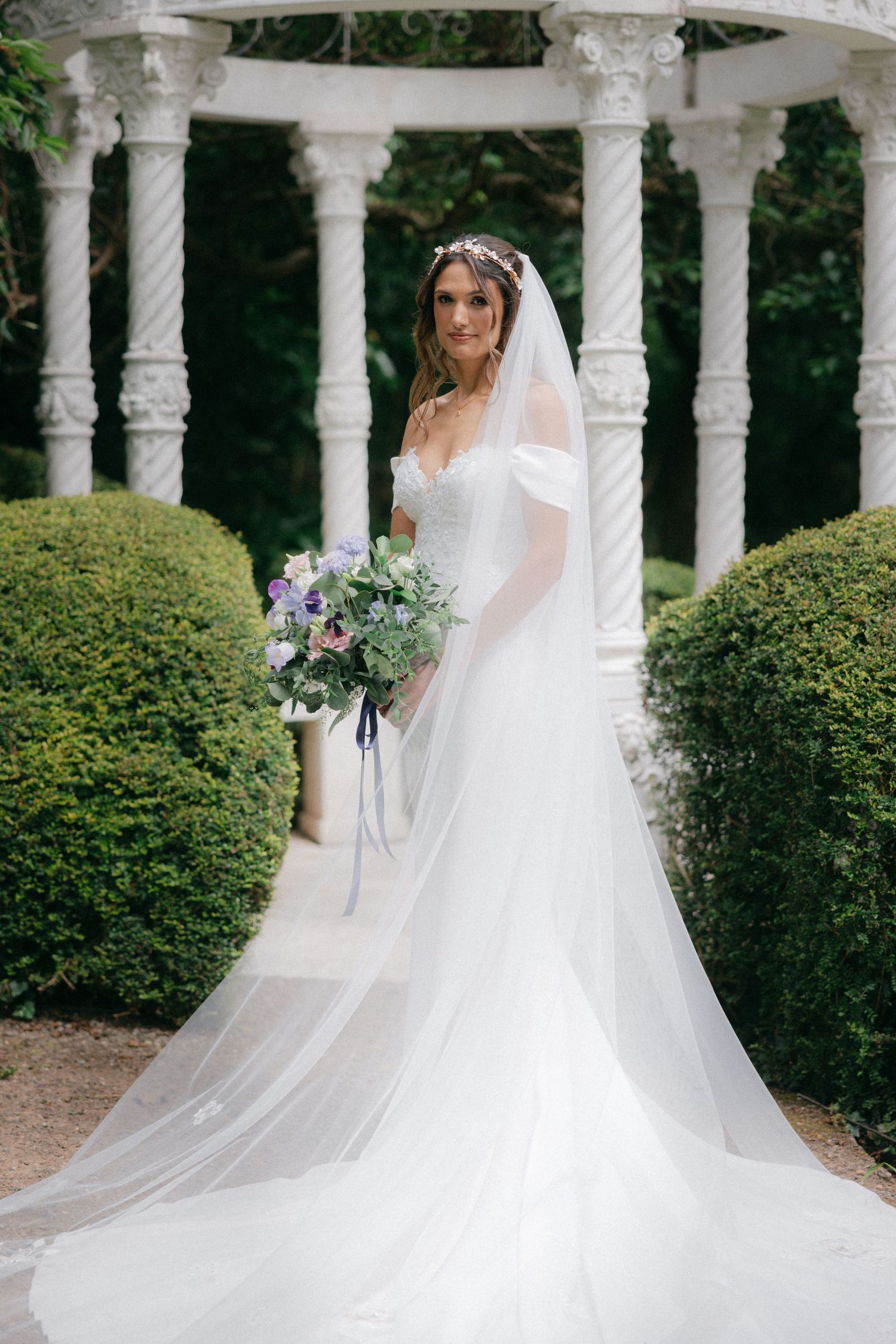 A flowing white wedding dress trails elegantly among garden pillars and greenery.