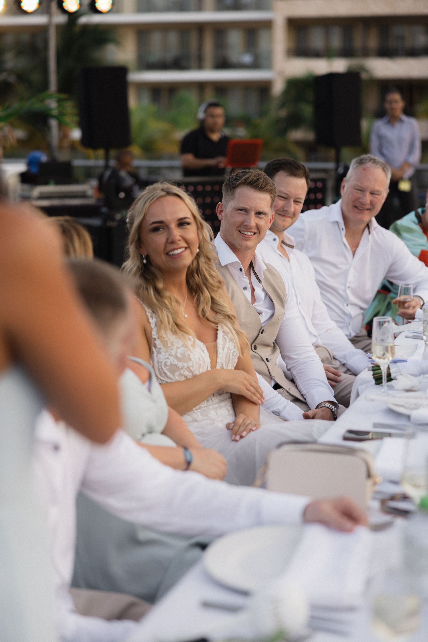 Wedding guests in white attire enjoying speeches at an outdoor reception.