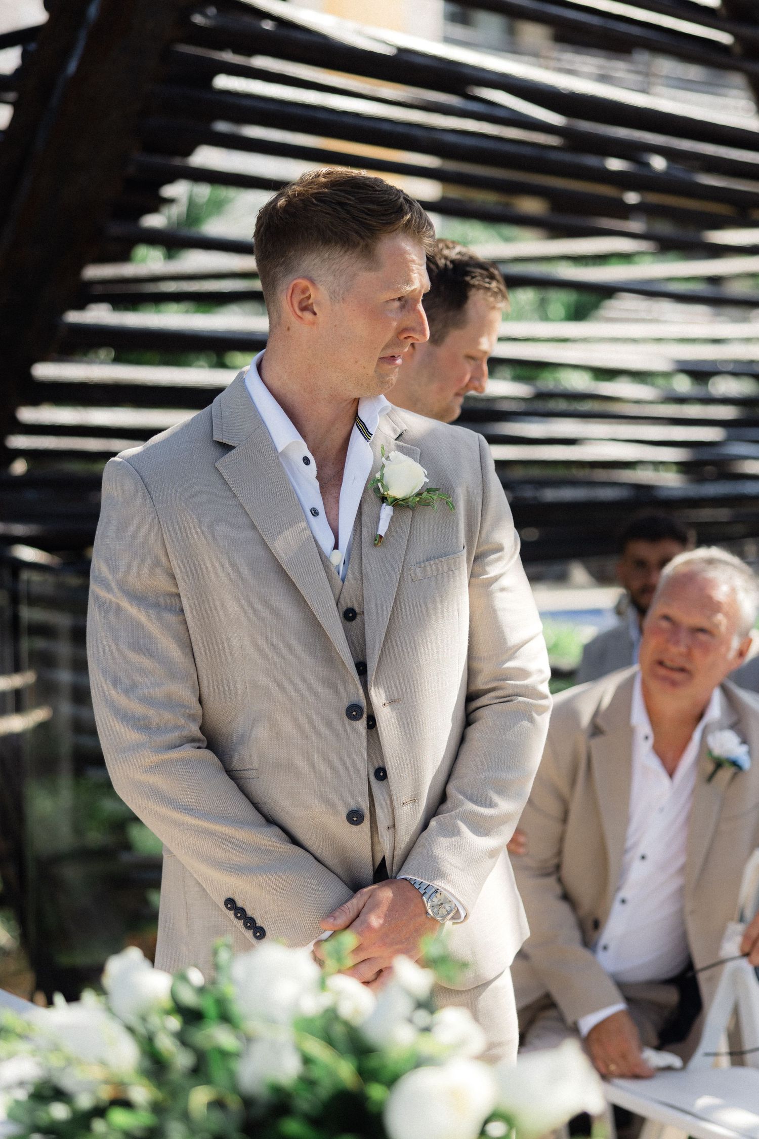 Wedding party members in beige suits gathered around floral arrangements outdoors.