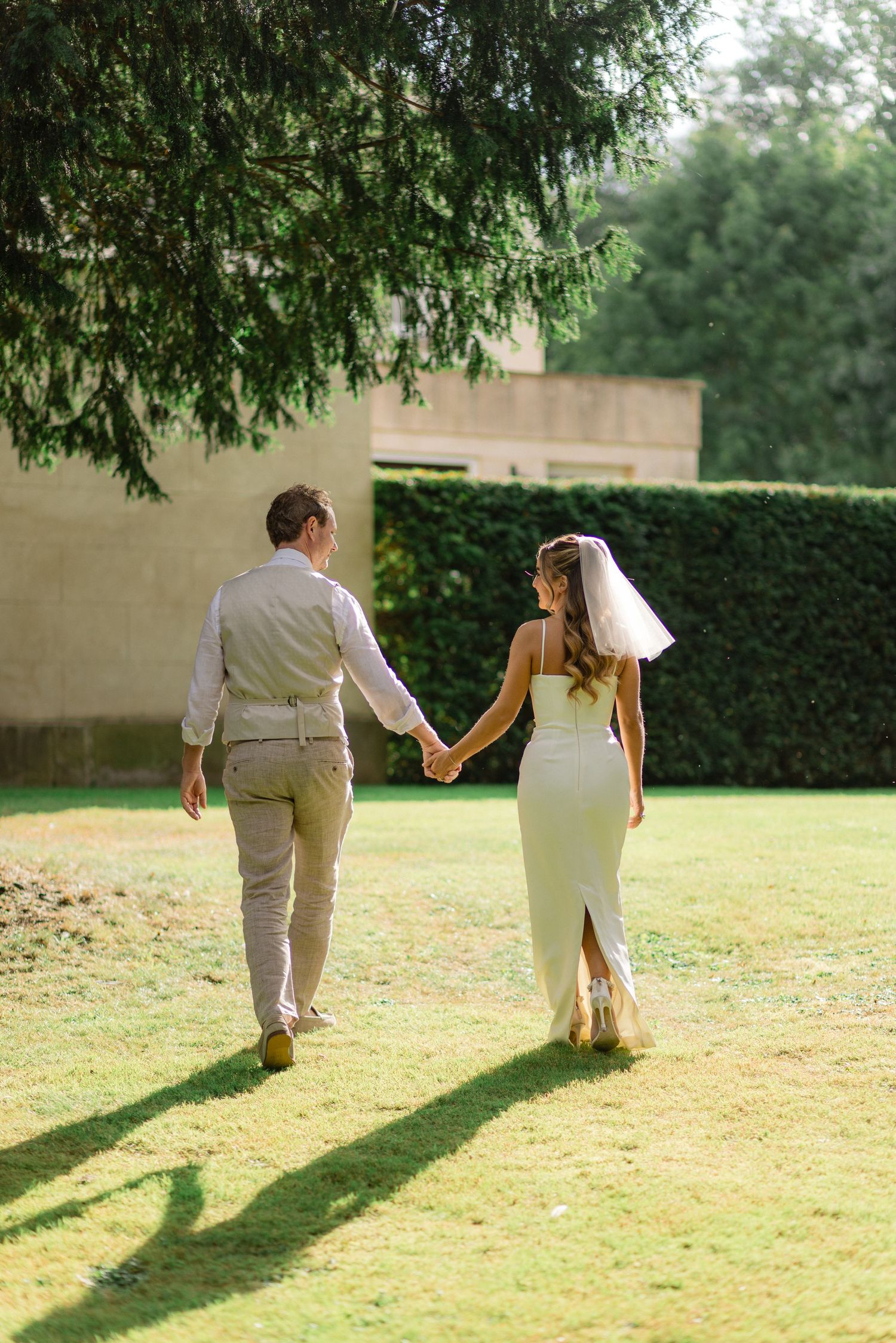 A romantic wedding couple walking hand in hand across a sunlit lawn, creating long shadows.