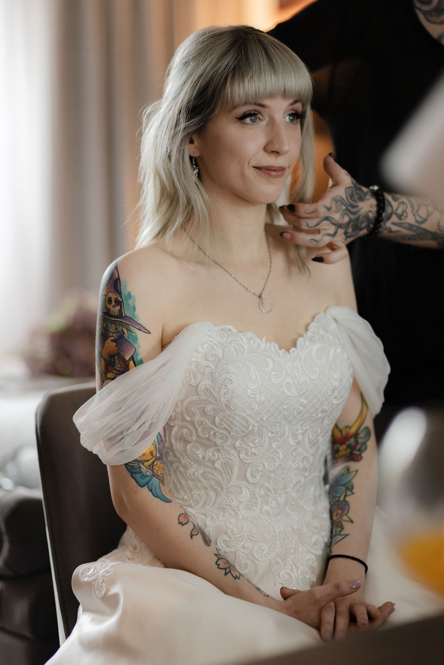 Bride in white lace dress showing colorful arm tattoos during wedding preparations.