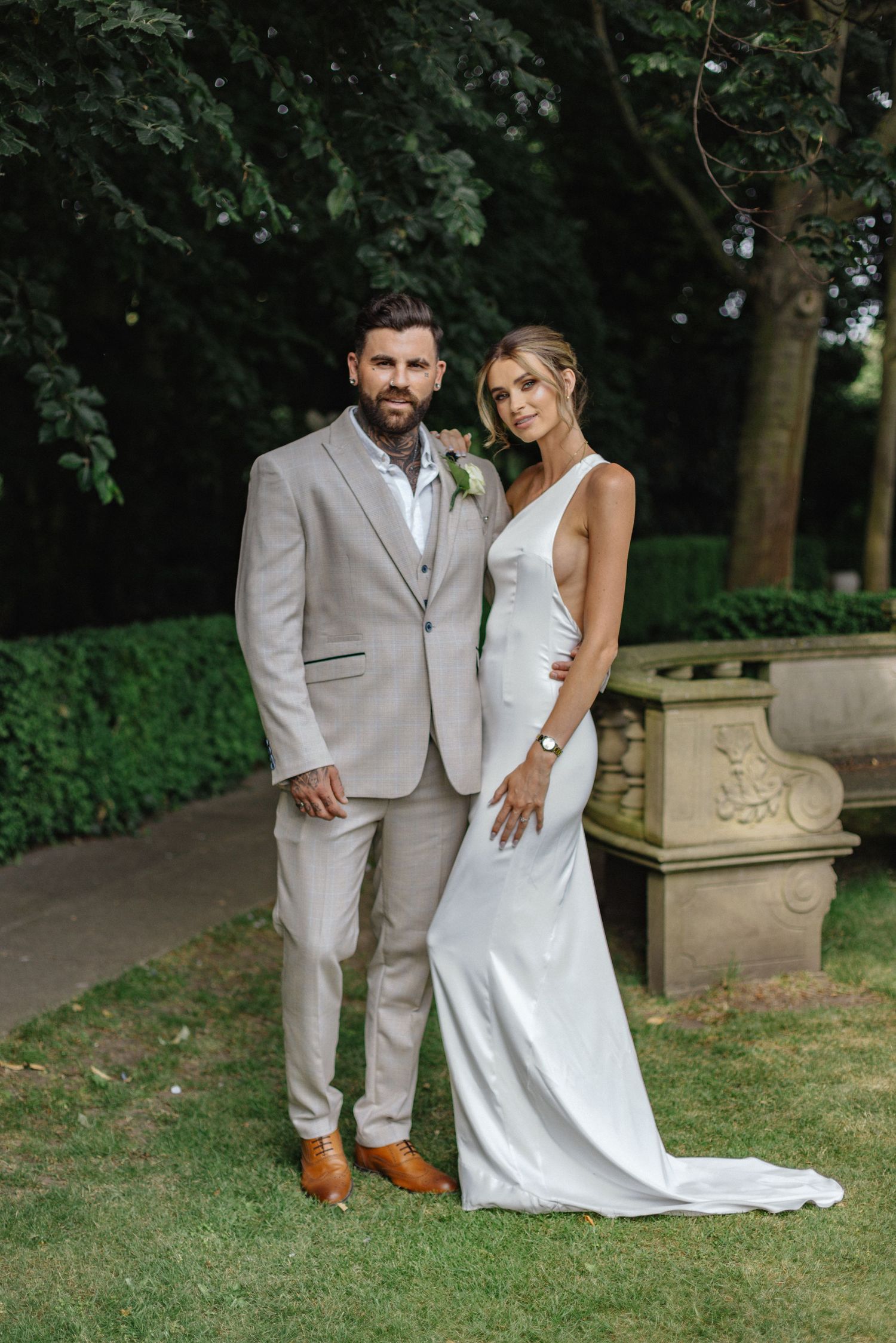 Wedding couple poses together on grassy lawn in elegant white and beige attire.