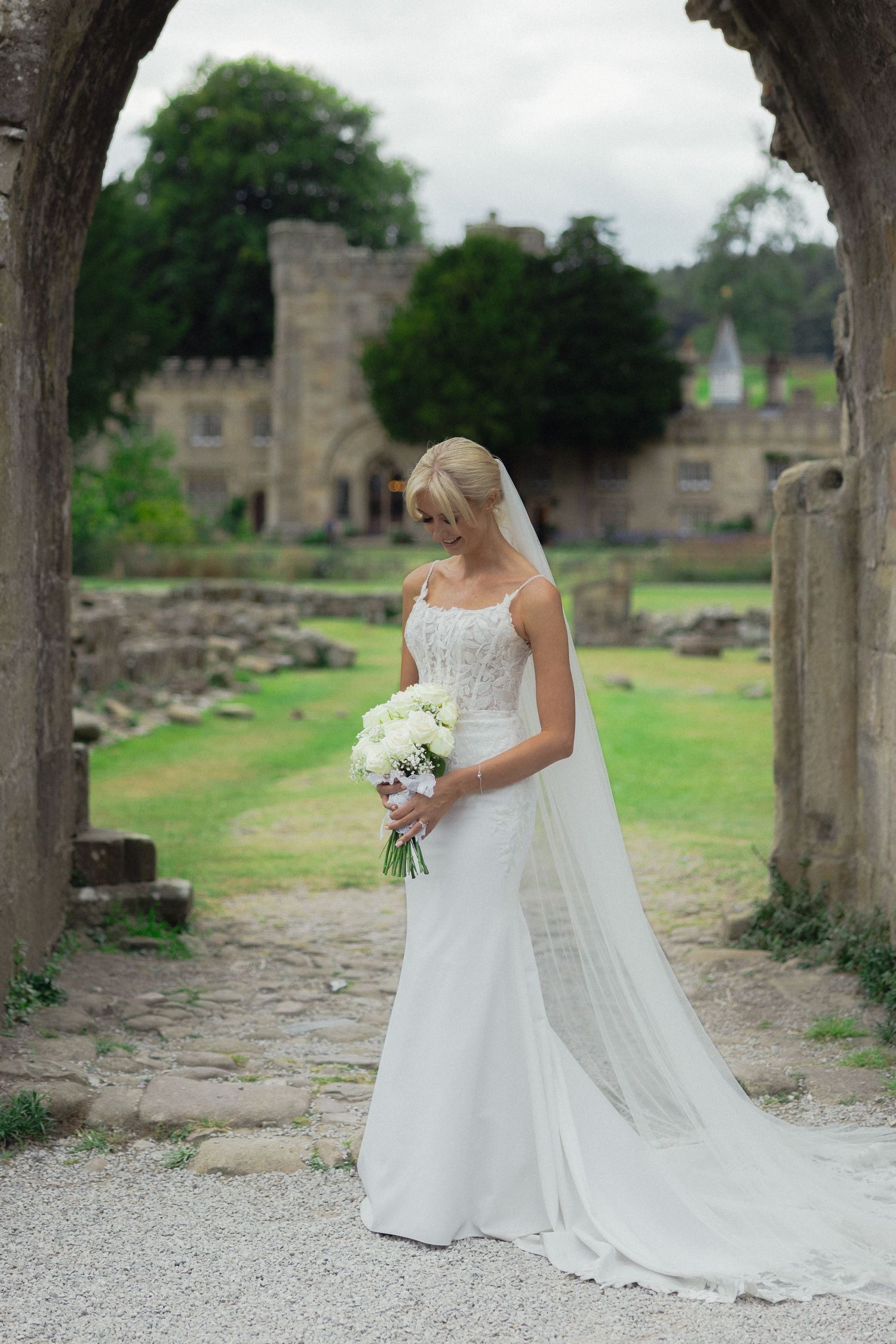 A bride in white gown holds bouquet while standing under historic stone arch with castle backdrop.