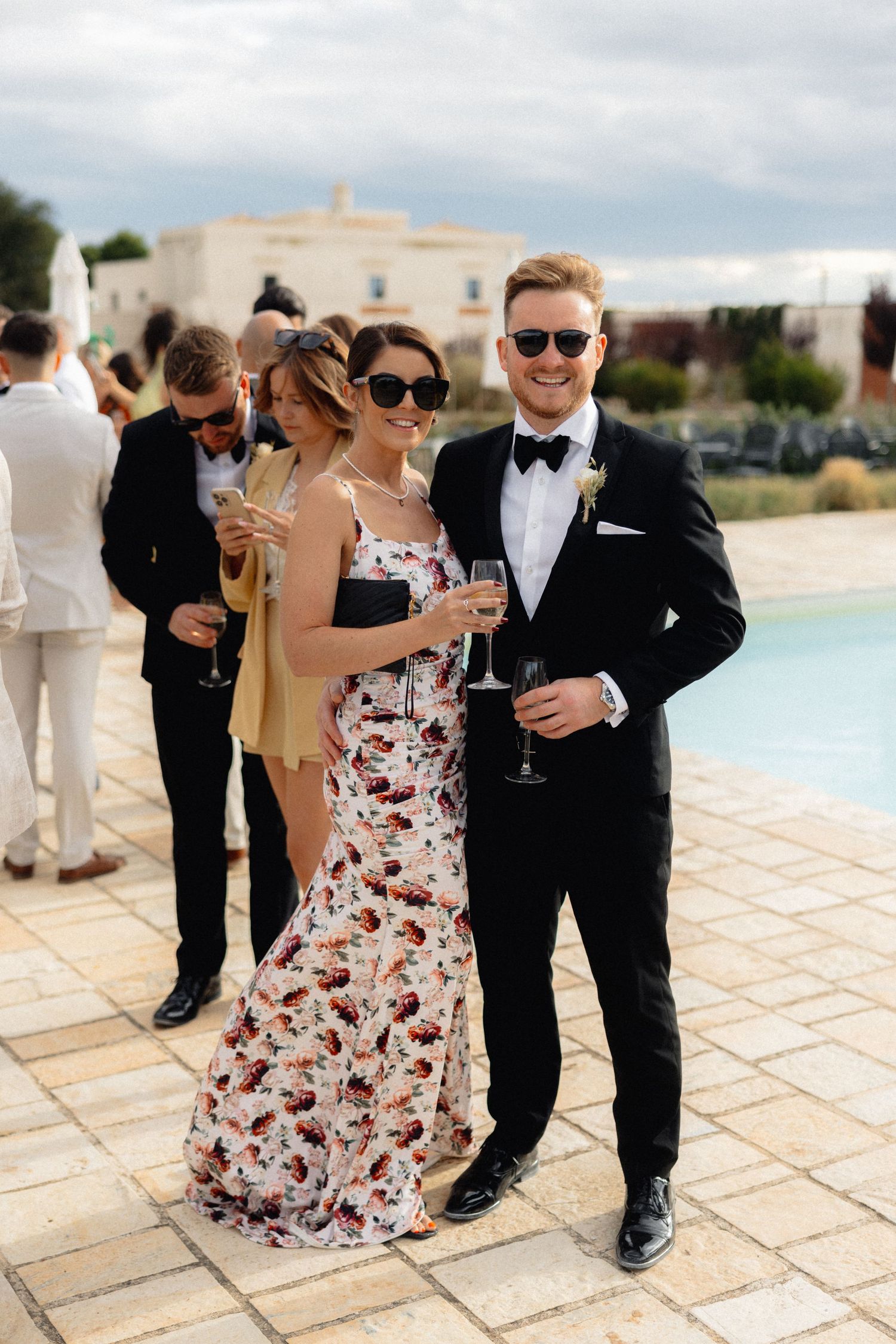Wedding guests in formal attire enjoying drinks and conversation by a poolside terrace.