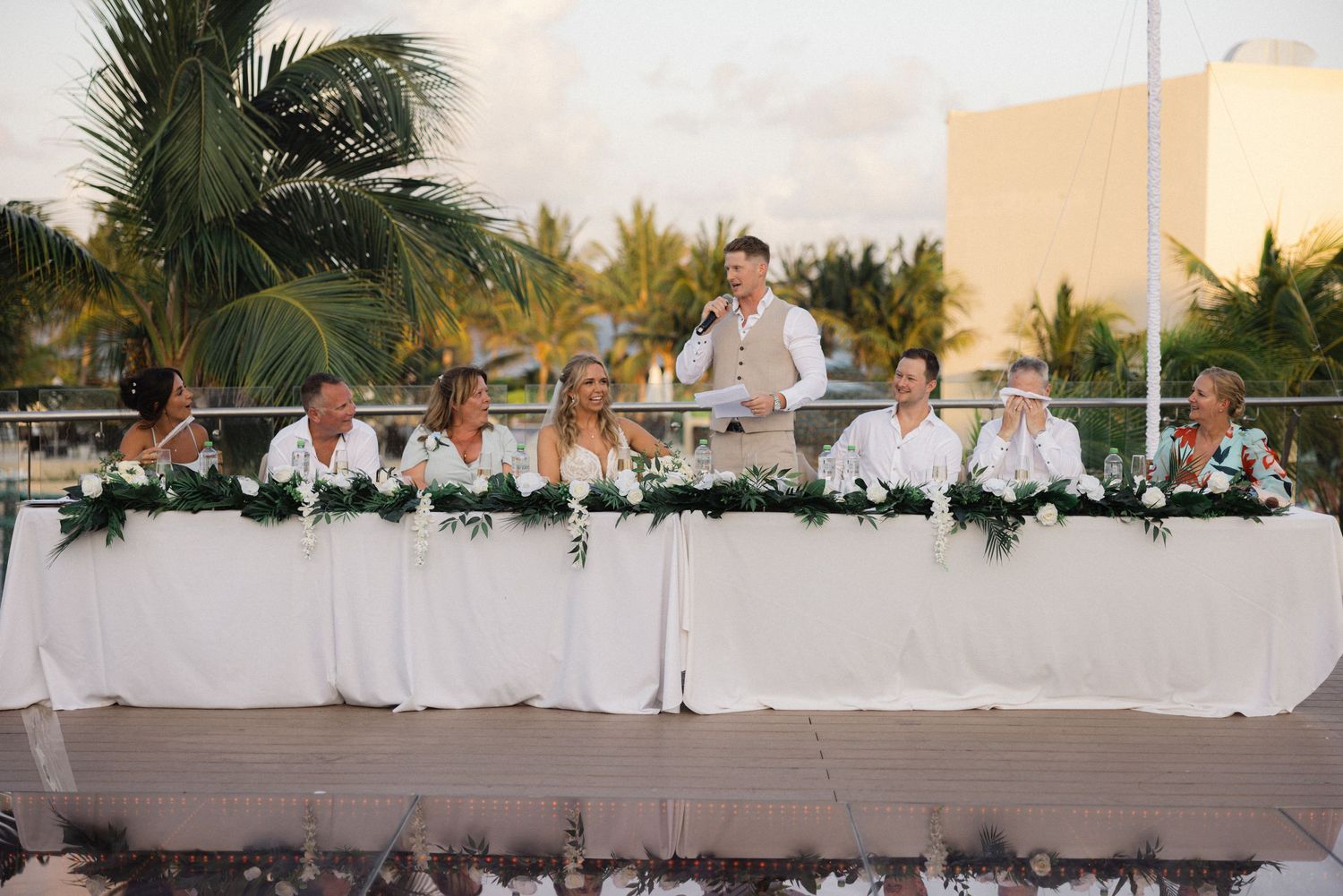 Wedding reception table setup at an outdoor tropical venue with palm trees and white decor.