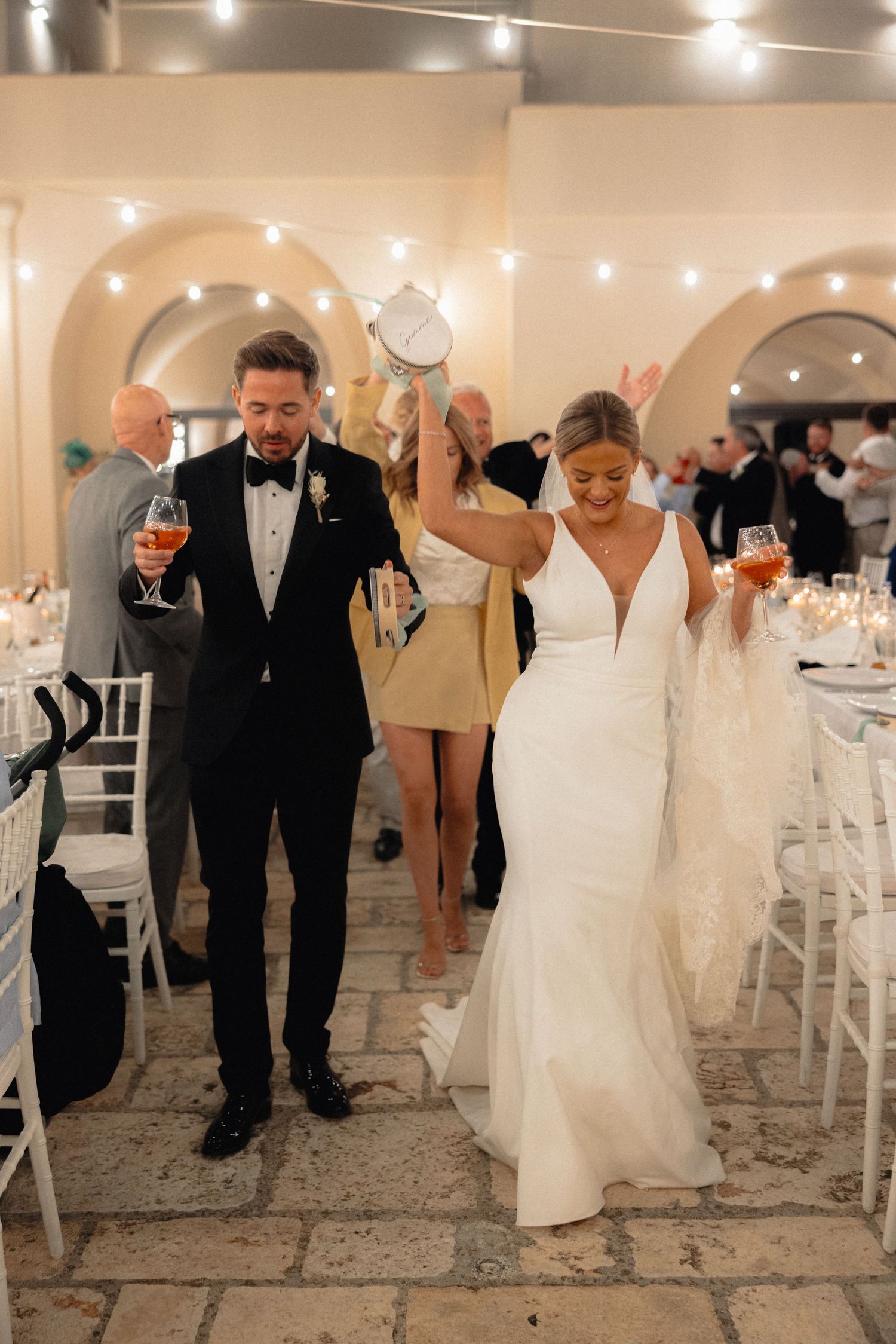 Wedding celebration with guests dancing in formal attire under string lights on a stone floor.