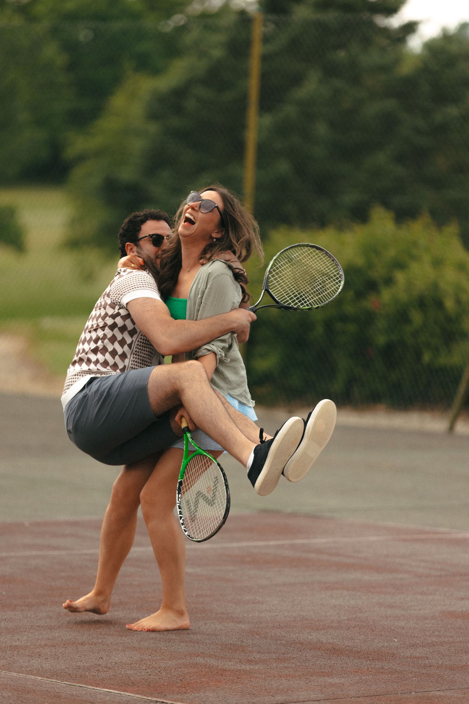 A couple playfully posing with tennis racquets on an outdoor court.