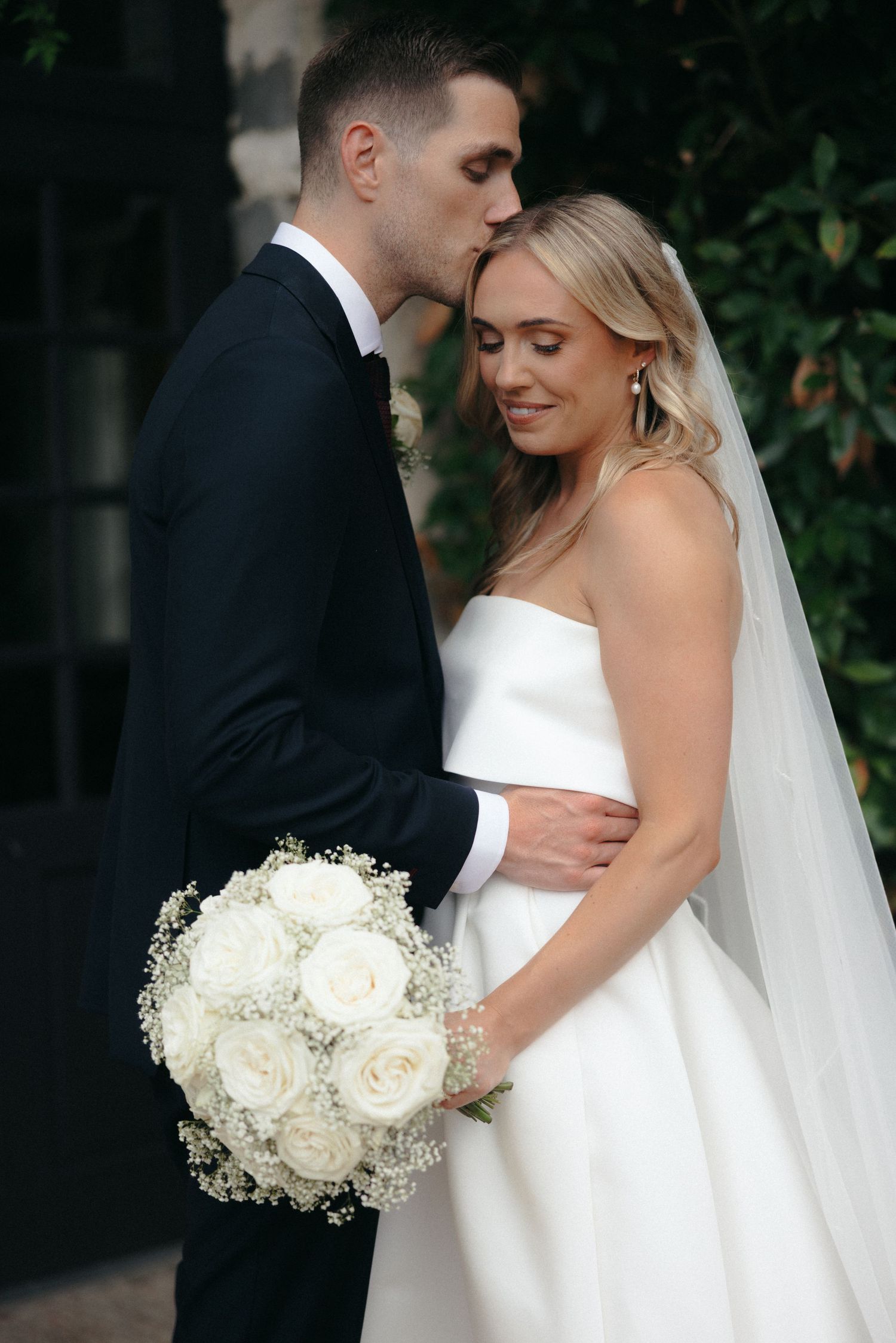 Romantic wedding portraits with white roses bouquet against garden backdrop.