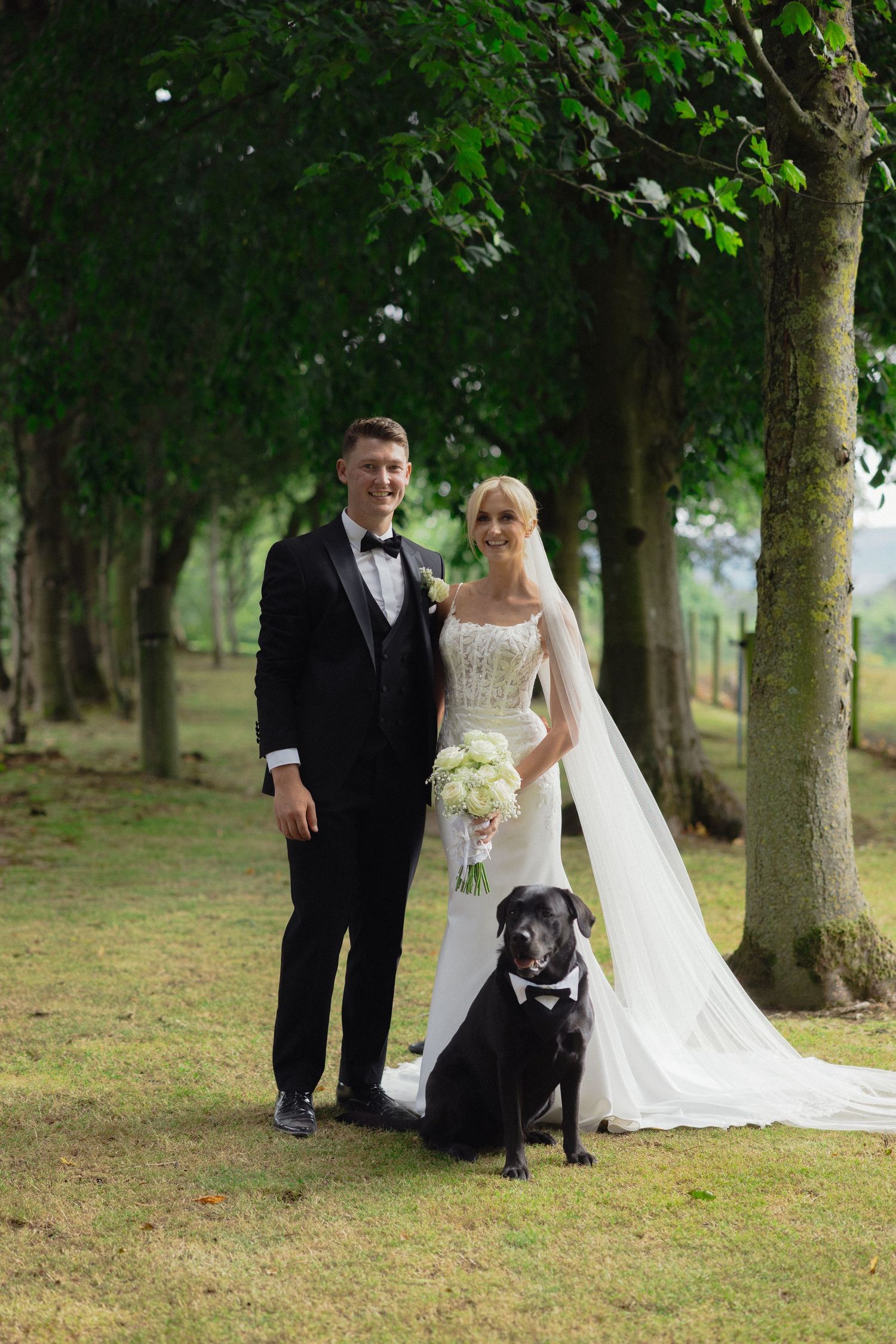 Wedding couple poses with their dog in wooded outdoor setting.