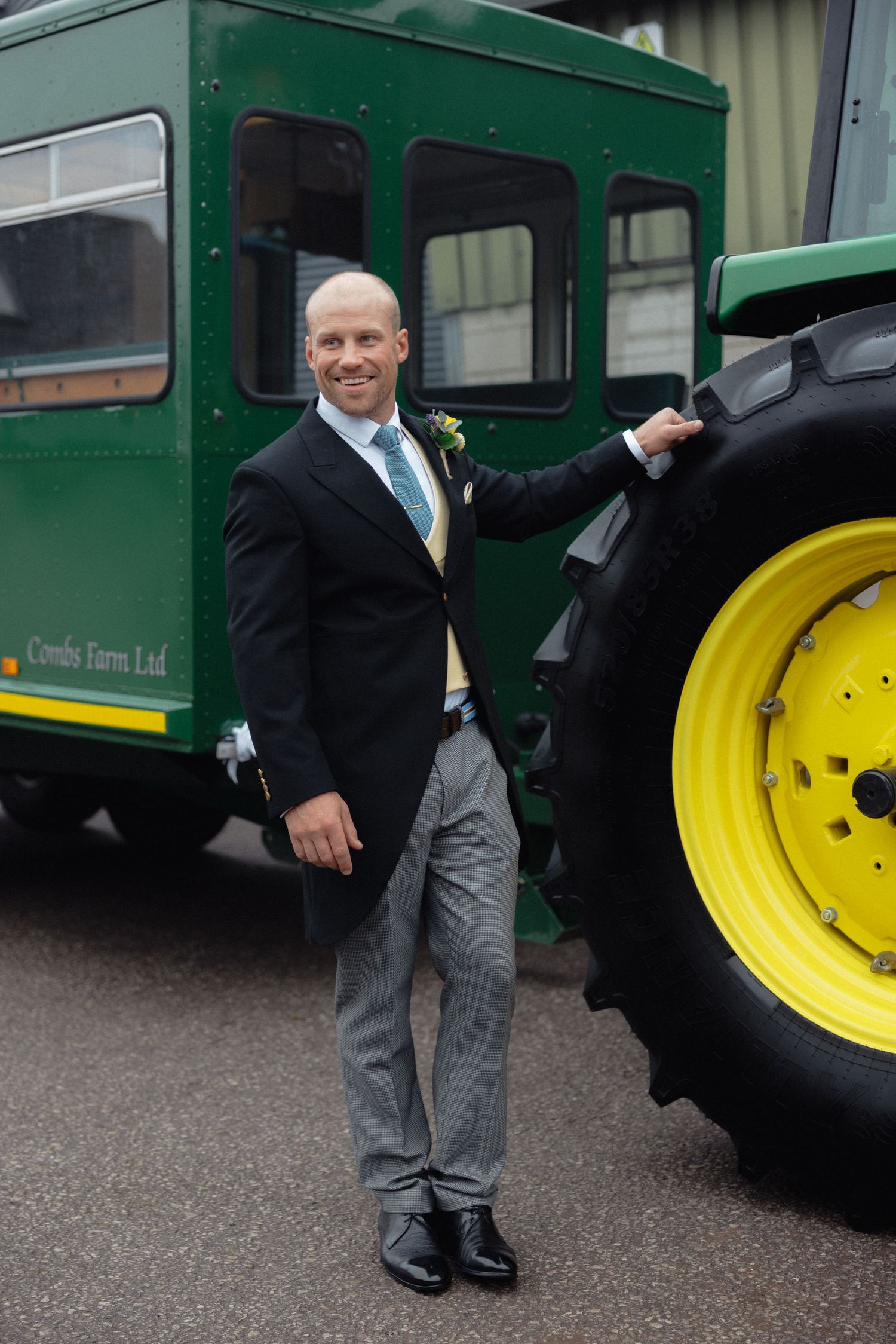 Person in formal wear posing playfully near a green tractor with yellow wheels.