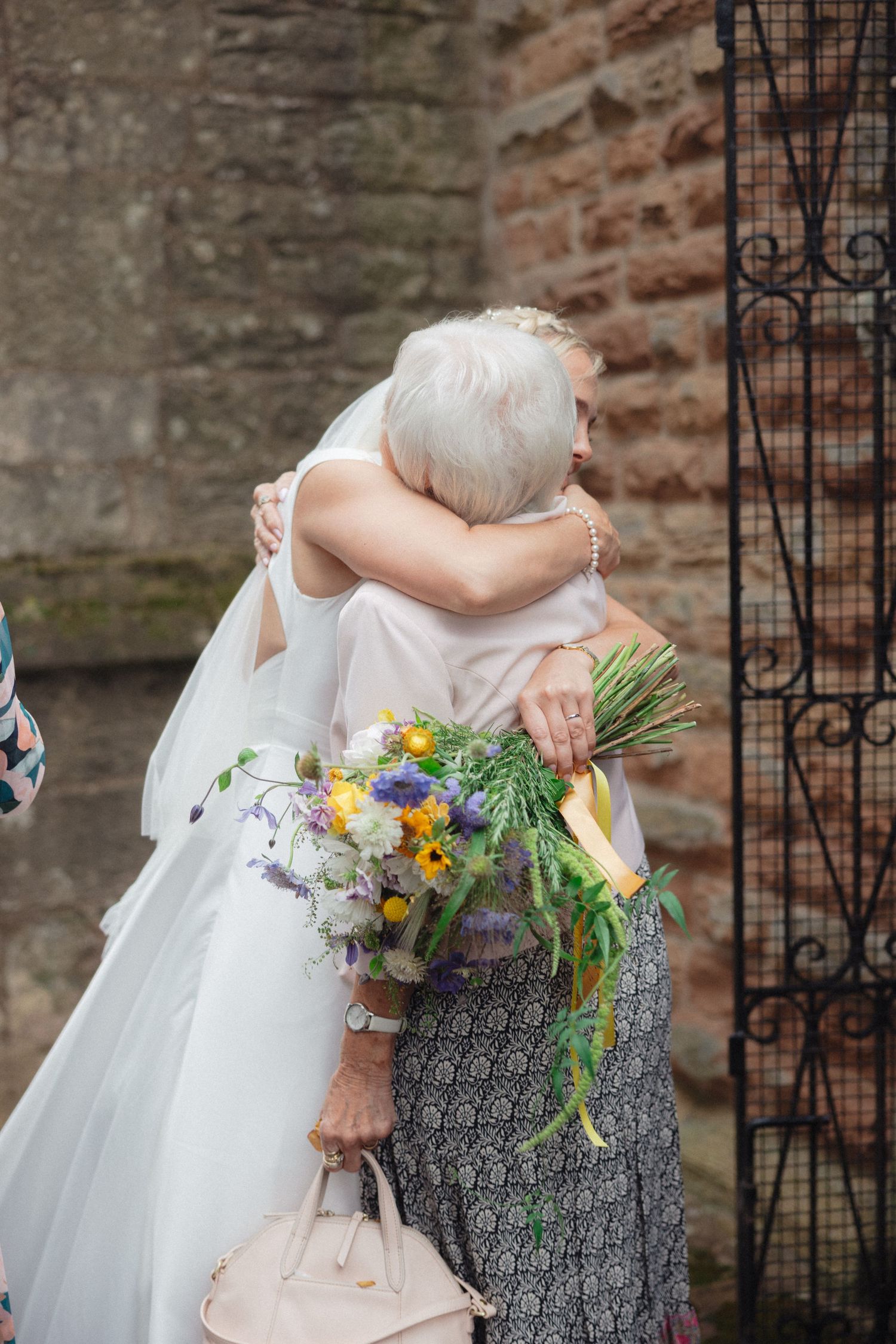 Bride in white dress hugging guest while holding colorful wildflower bouquet.