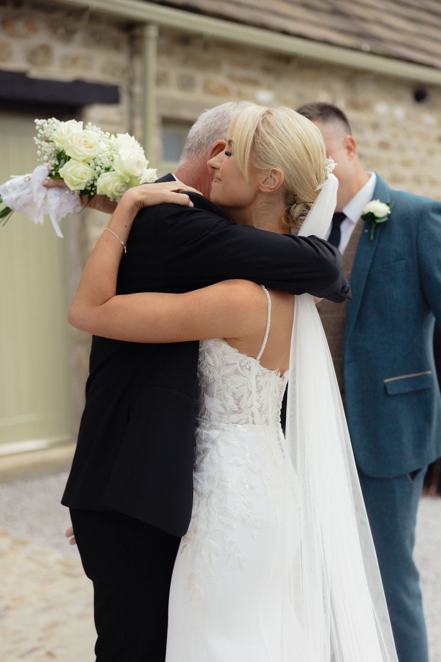 Wedding party members in formal attire embrace during an emotional moment at an outdoor venue.