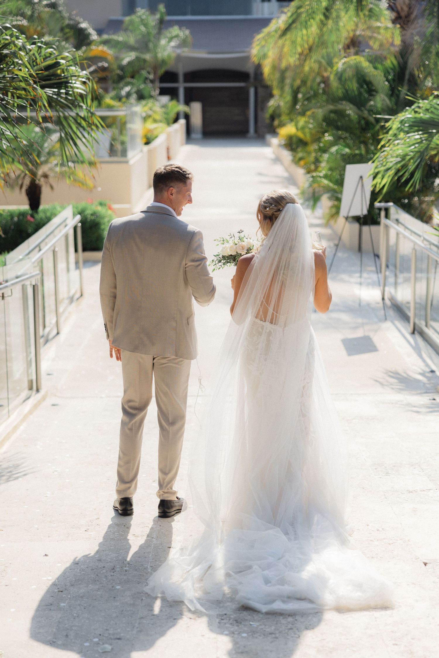 Couple walks away hand in hand on white pathway surrounded by lush tropical landscaping.
