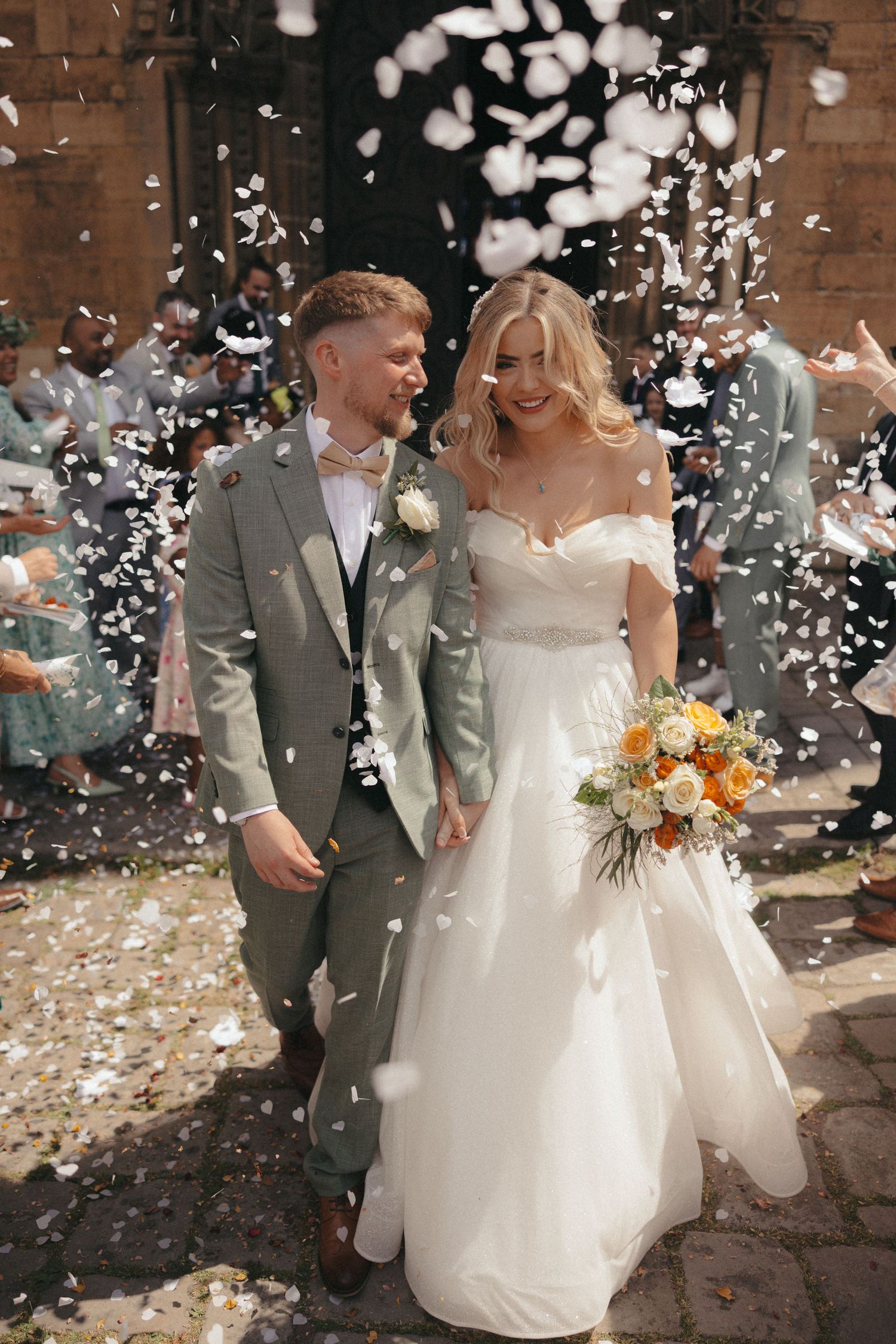 A wedding couple walking through a shower of white confetti outside a brick building.