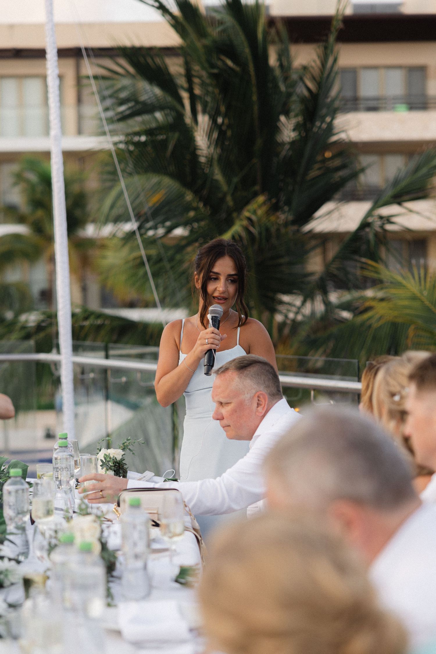 Outdoor wedding reception with guests listening to speeches under palm trees.