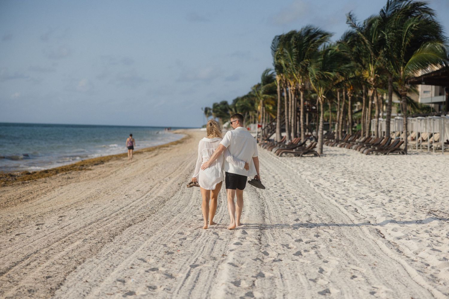 A couple strolls together on a scenic beach with palm trees and turquoise waters in the background.